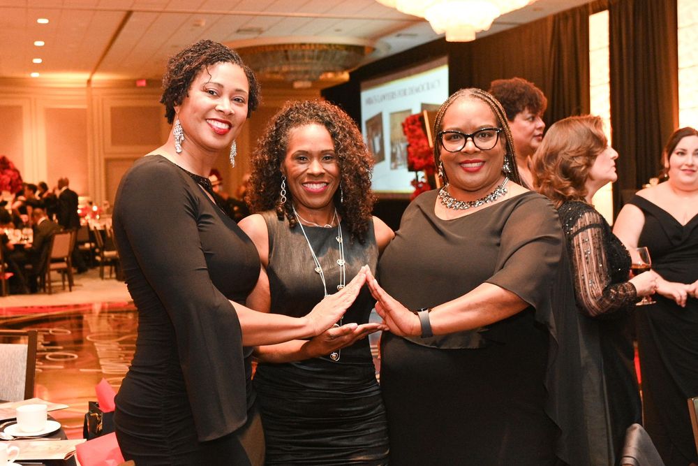 Three people pose together at an indoor formal event wearing elegant black attire.