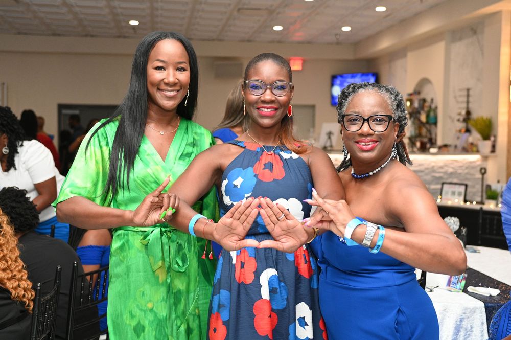 Three women in vibrant dresses of green, blue and royal blue pose together at a social event indoors.