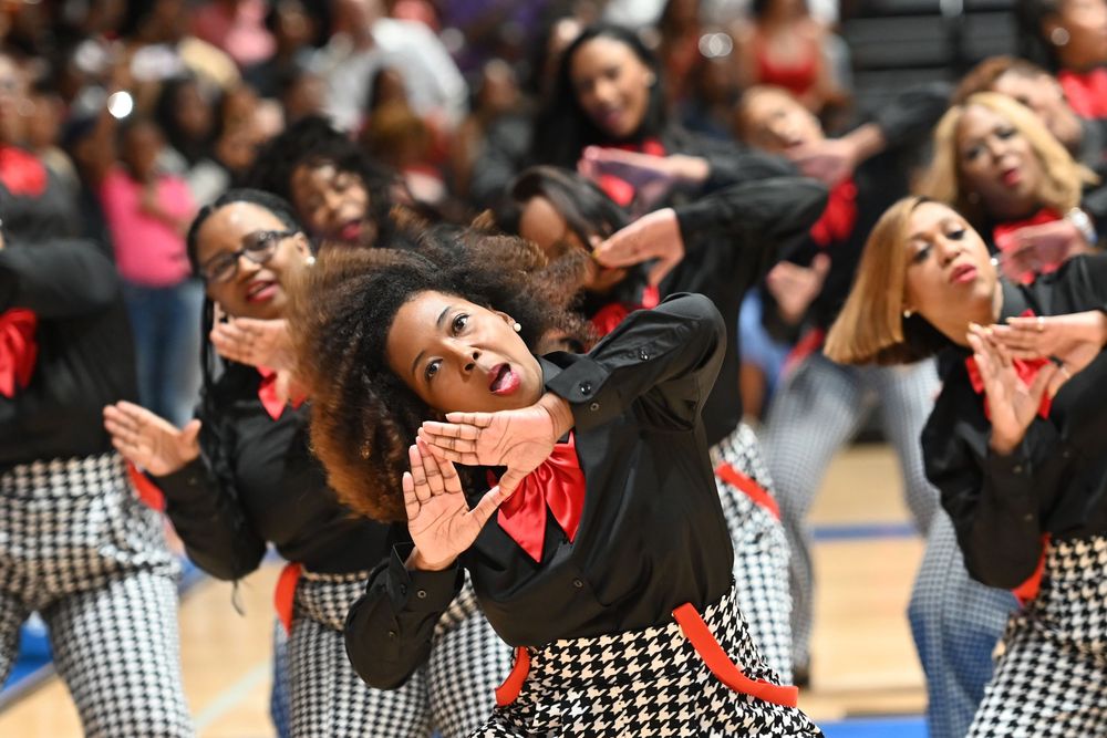 Dancers in matching black and white houndstooth outfits perform an energetic dance routine at an indoor venue.