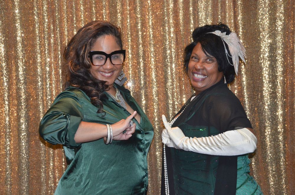 Two people in green satin jackets and white gloves pose in front of a sparkly gold backdrop.