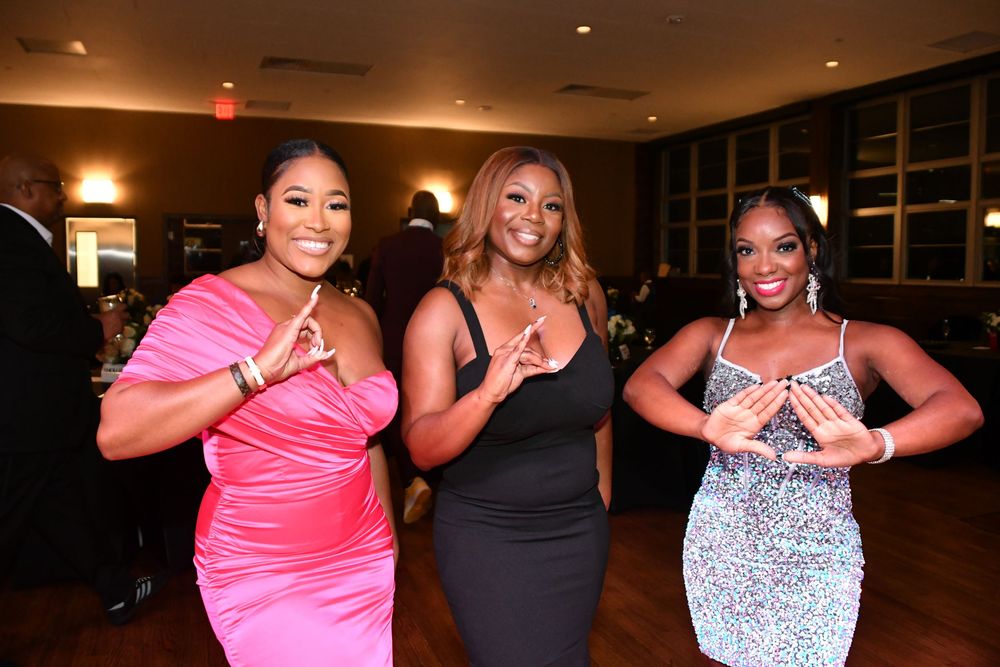 Three women in glamorous evening dresses pose together at an indoor event with warm lighting.