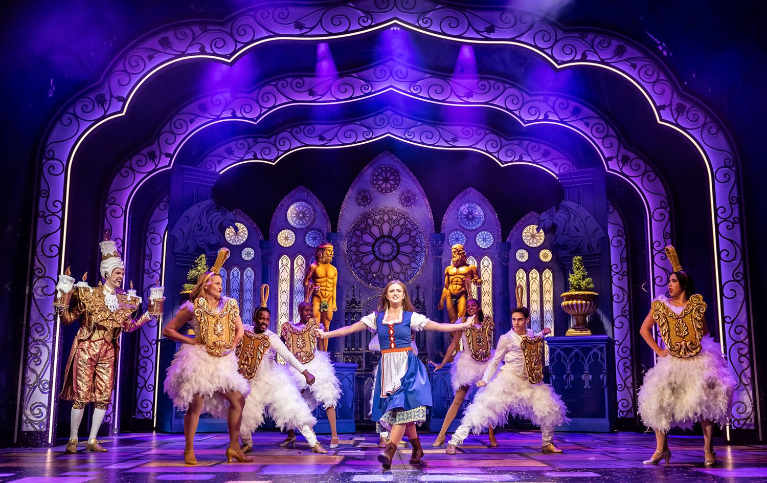 Theatrical stage production featuring elaborate purple-lit archways and performers in ornate costumes with feathered props.