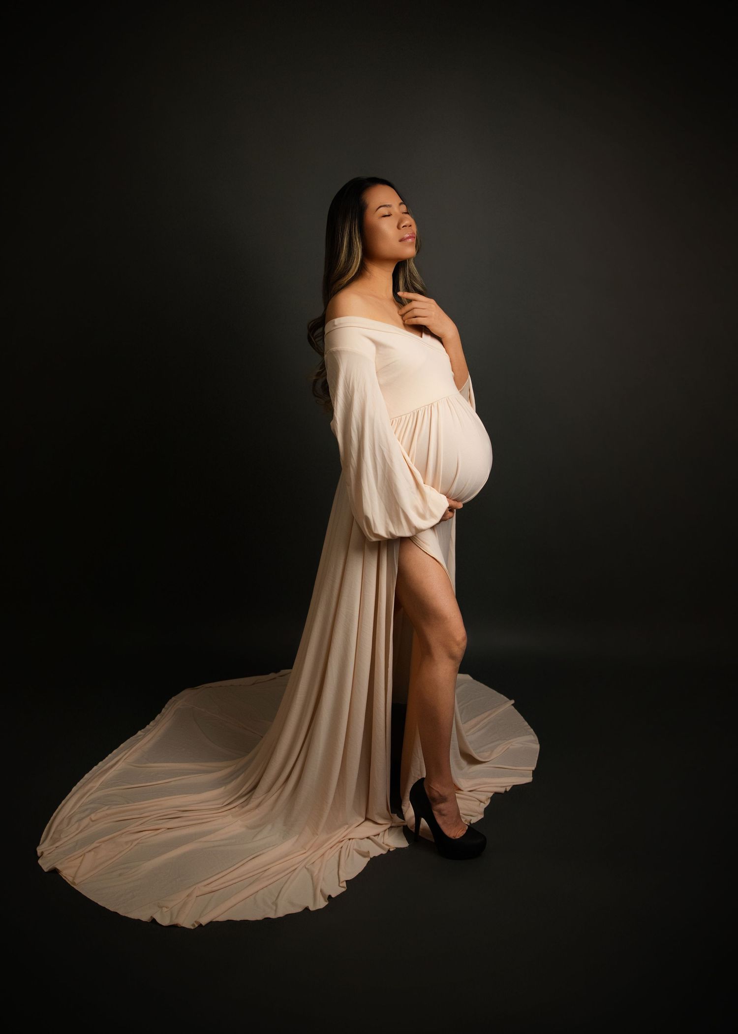 Studio maternity portrait featuring an elegant off-shoulder white flowing gown with long train against a dark backdrop.