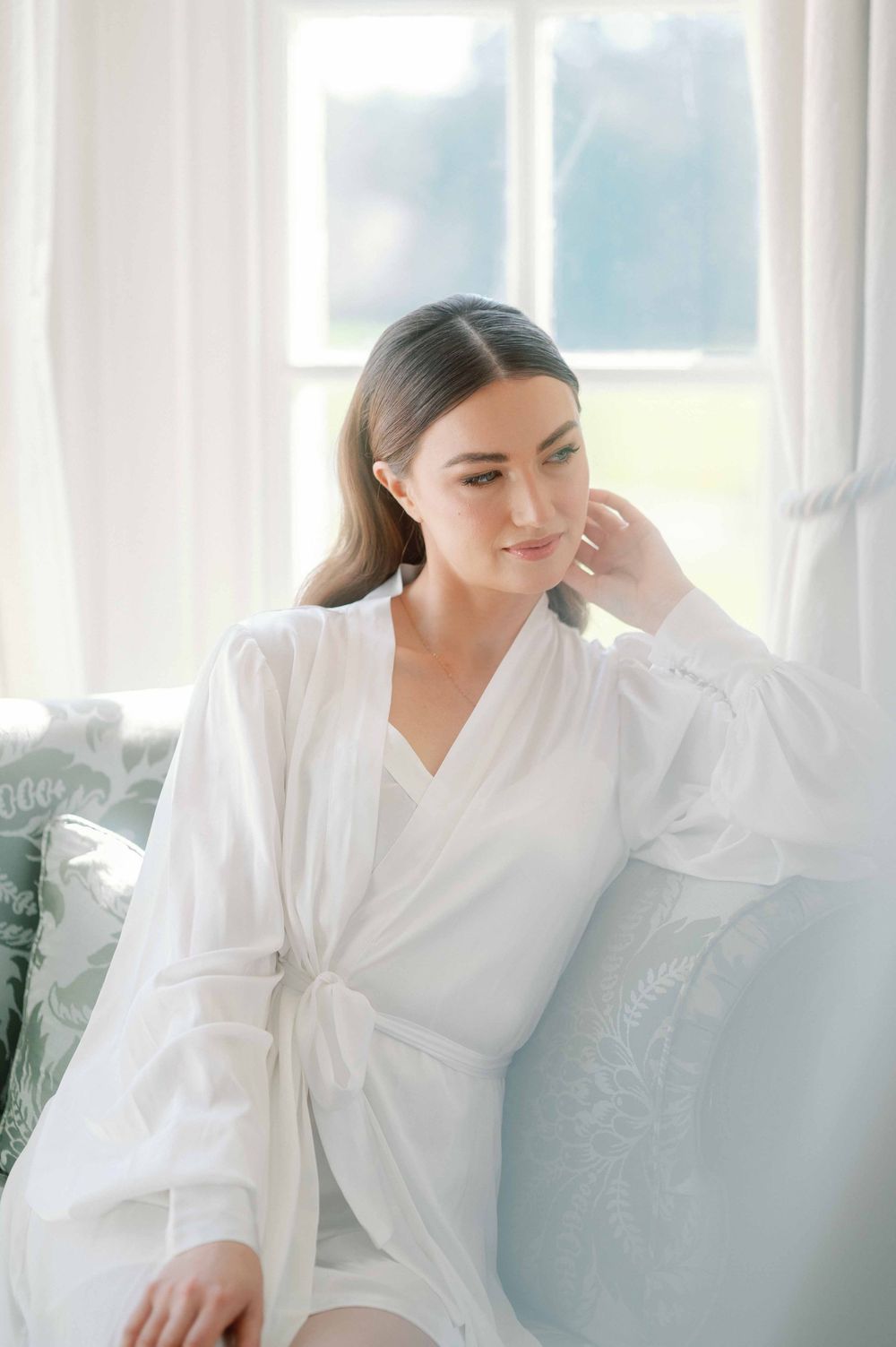 Woman in white silk robe sitting by window with soft natural light in elegant boudoir setting.