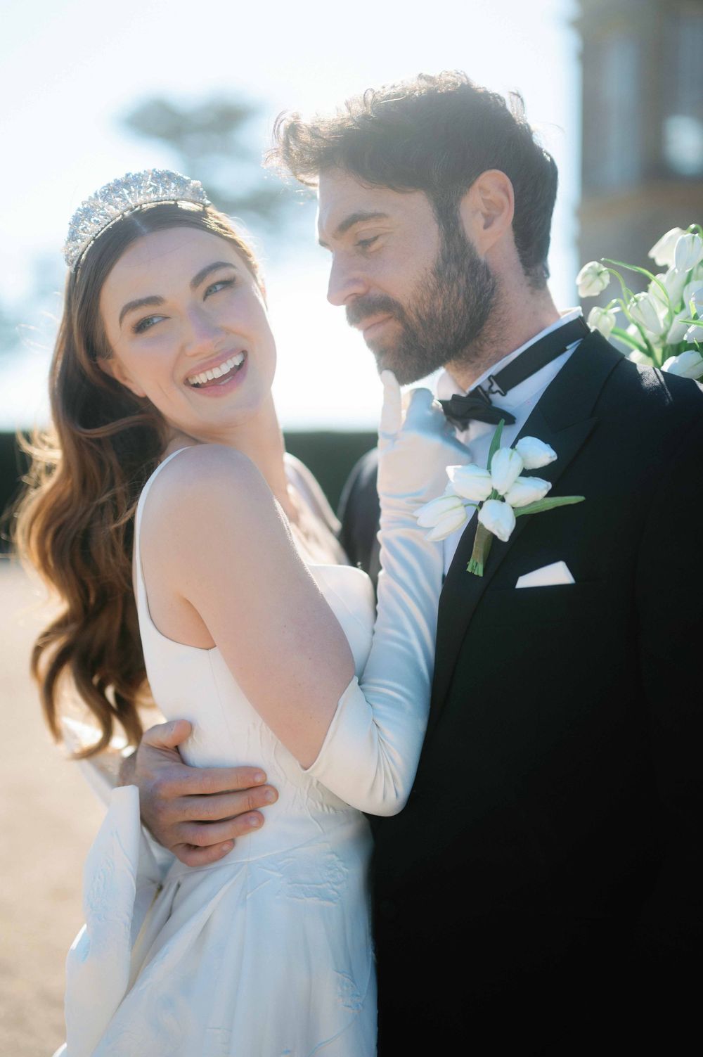 A joyful wedding couple embraces in soft sunlight while wearing elegant white dress and black tuxedo with bow tie.