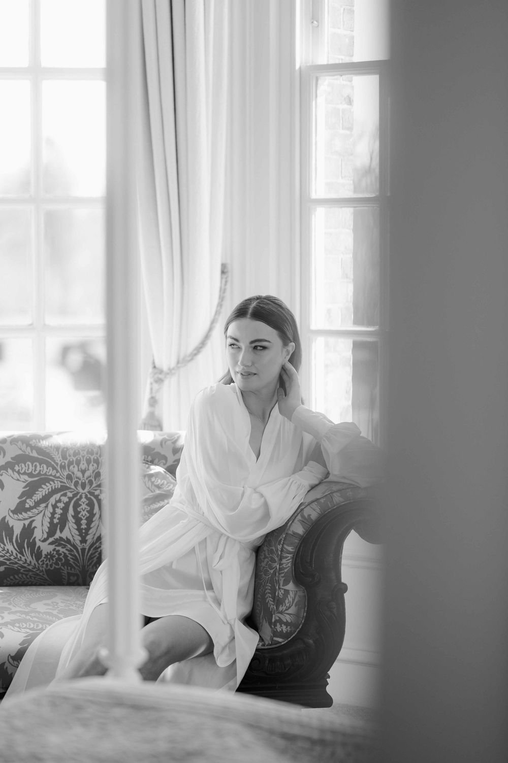 A black and white boudoir photograph shows a person in a white robe sitting on an elegant couch near bright windows.