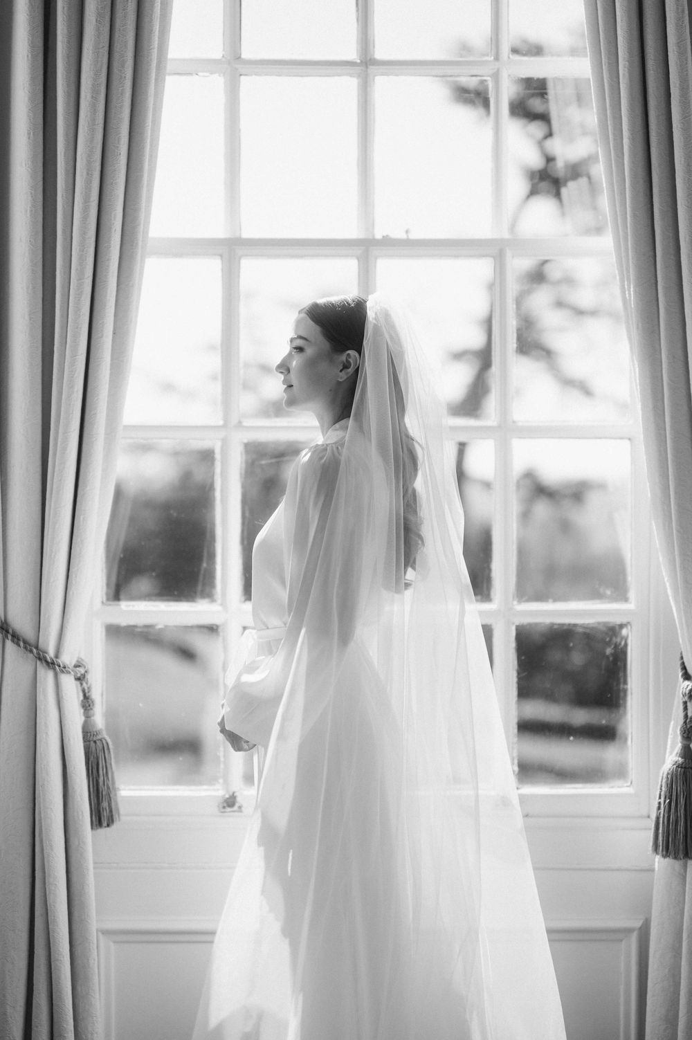 A bride in a flowing white veil stands silhouetted by bright window light in an elegant room with floor-length curtains.