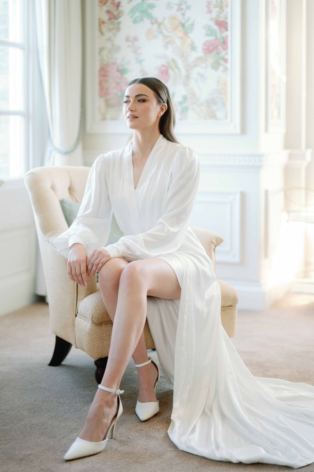 Elegant bridal scene with flowing white robe and heels in a bright room with floral wallpaper and cream armchair.