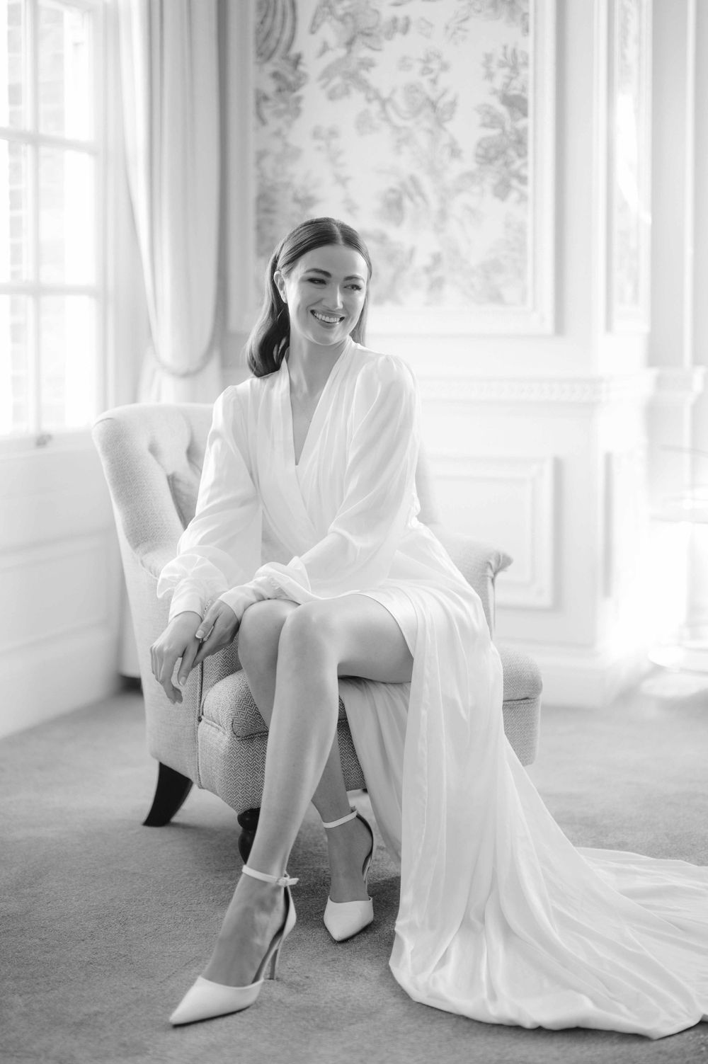 A person in a white robe sits elegantly on a light-colored chair against a sunlit wall with floral wallpaper.