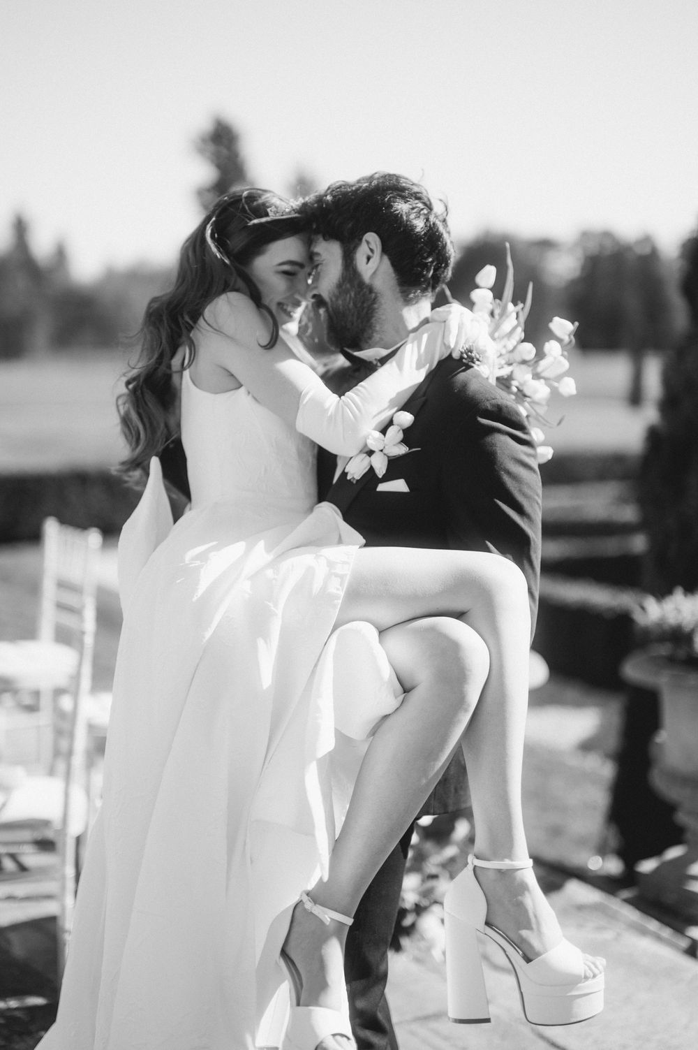 Black and white wedding photo showing a romantic outdoor moment with bride in platform shoes and flowing dress.