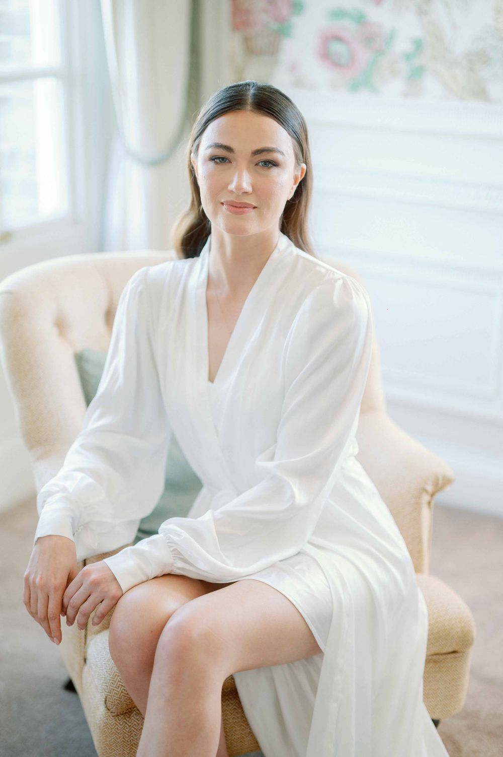 Person wearing white robe sitting elegantly in beige armchair by window in bright minimal room setting.