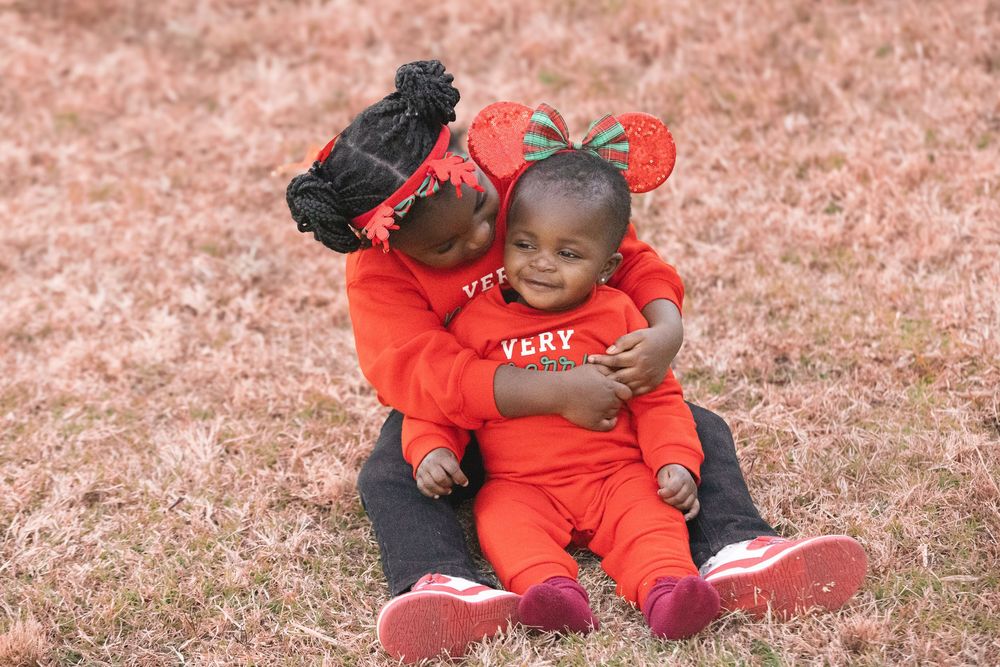 Two young children in matching red outfits sit on grass, with the older child hugging and kissing the baby.