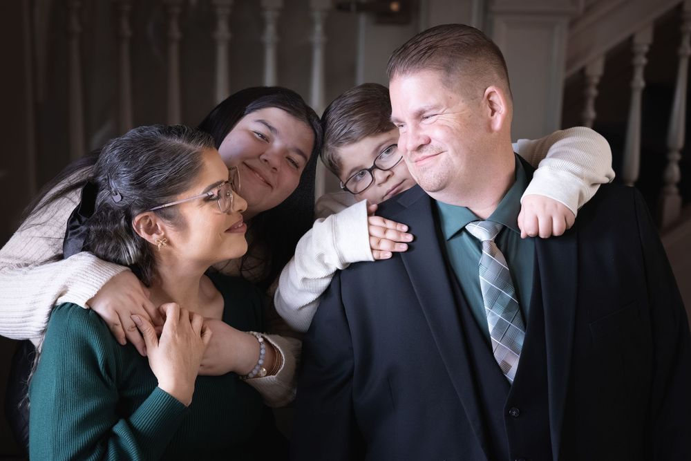 Family group hug with man in suit and green tie smiling as three people embrace him warmly indoors.