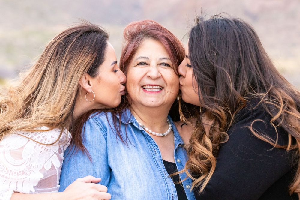Two daughters kissing their smiling mother on the cheeks in a warm family portrait outdoors.