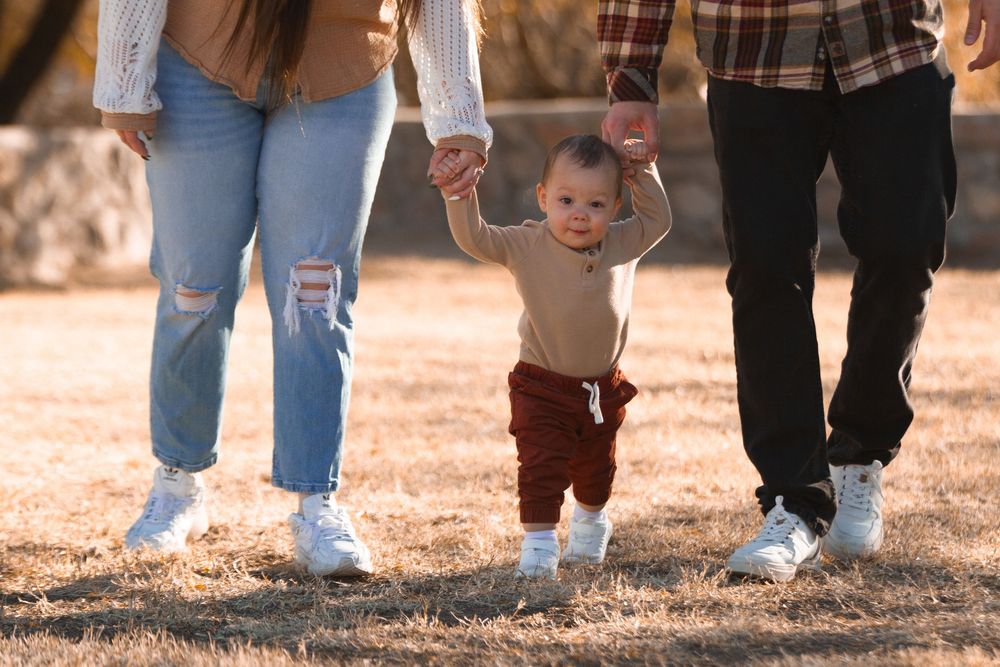 Parents holding hands with their baby learning to walk outdoors in a sunny park setting.