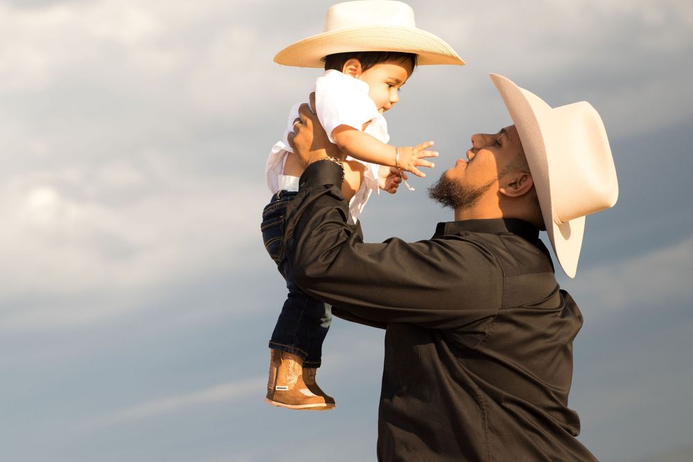 A father in a white cowboy hat lifts his young child wearing a matching hat against a cloudy sky.