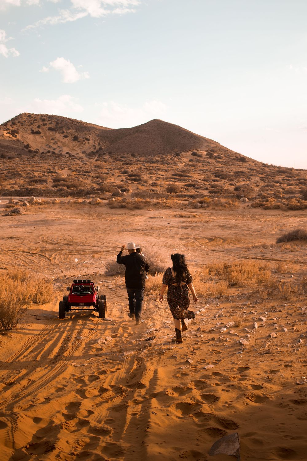 Two people walking toward a red dune buggy in a vast desert landscape with sandy terrain and a hill in the background.
