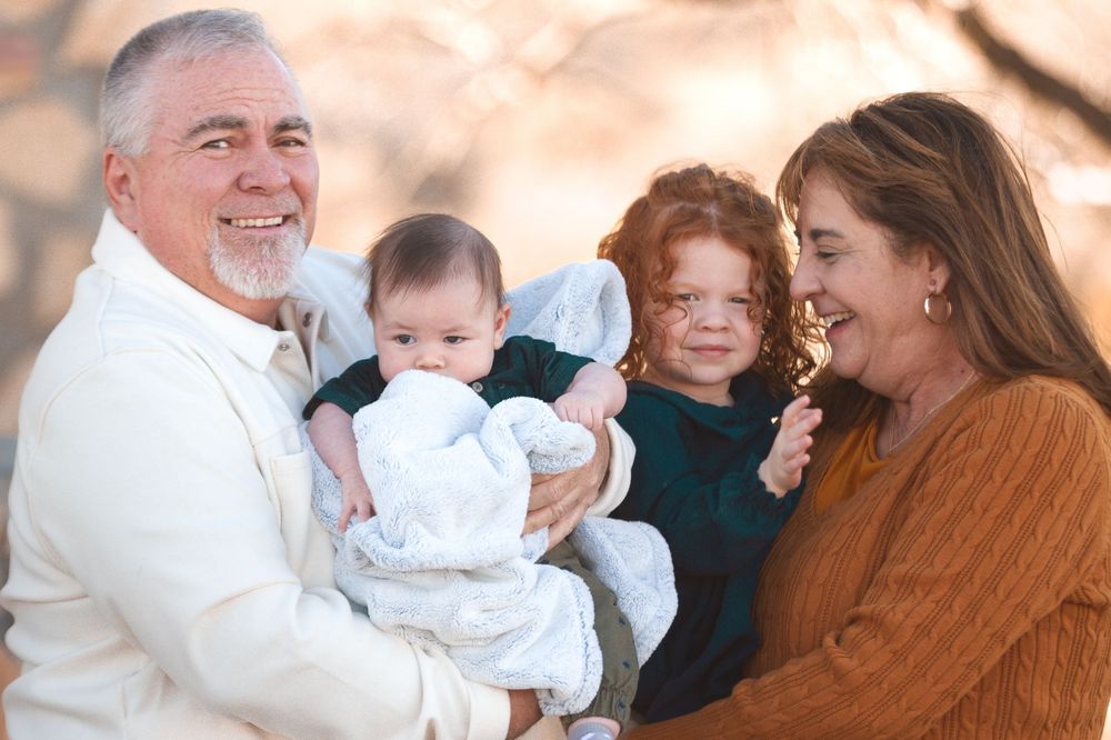 Grandparents smiling and holding two young grandchildren, a baby and a toddler, outdoors in warm autumn light.