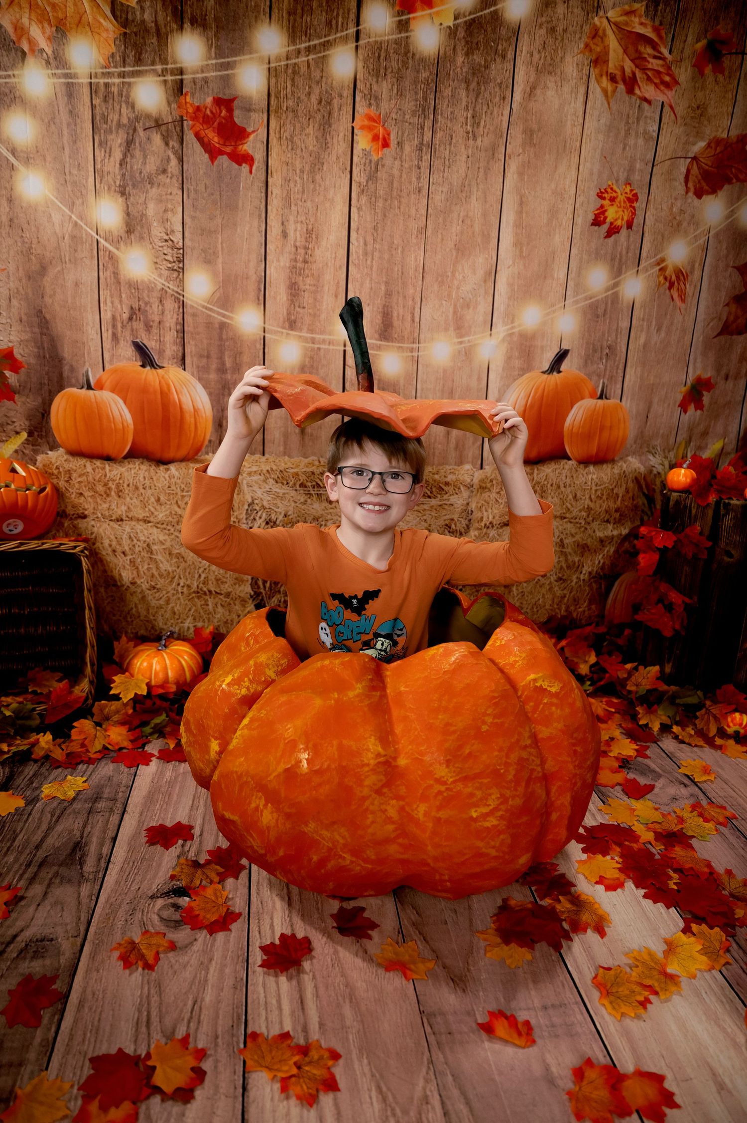 A festive autumn photo setup with a large orange pumpkin, string lights, fall leaves, and wooden backdrop for Halloween.