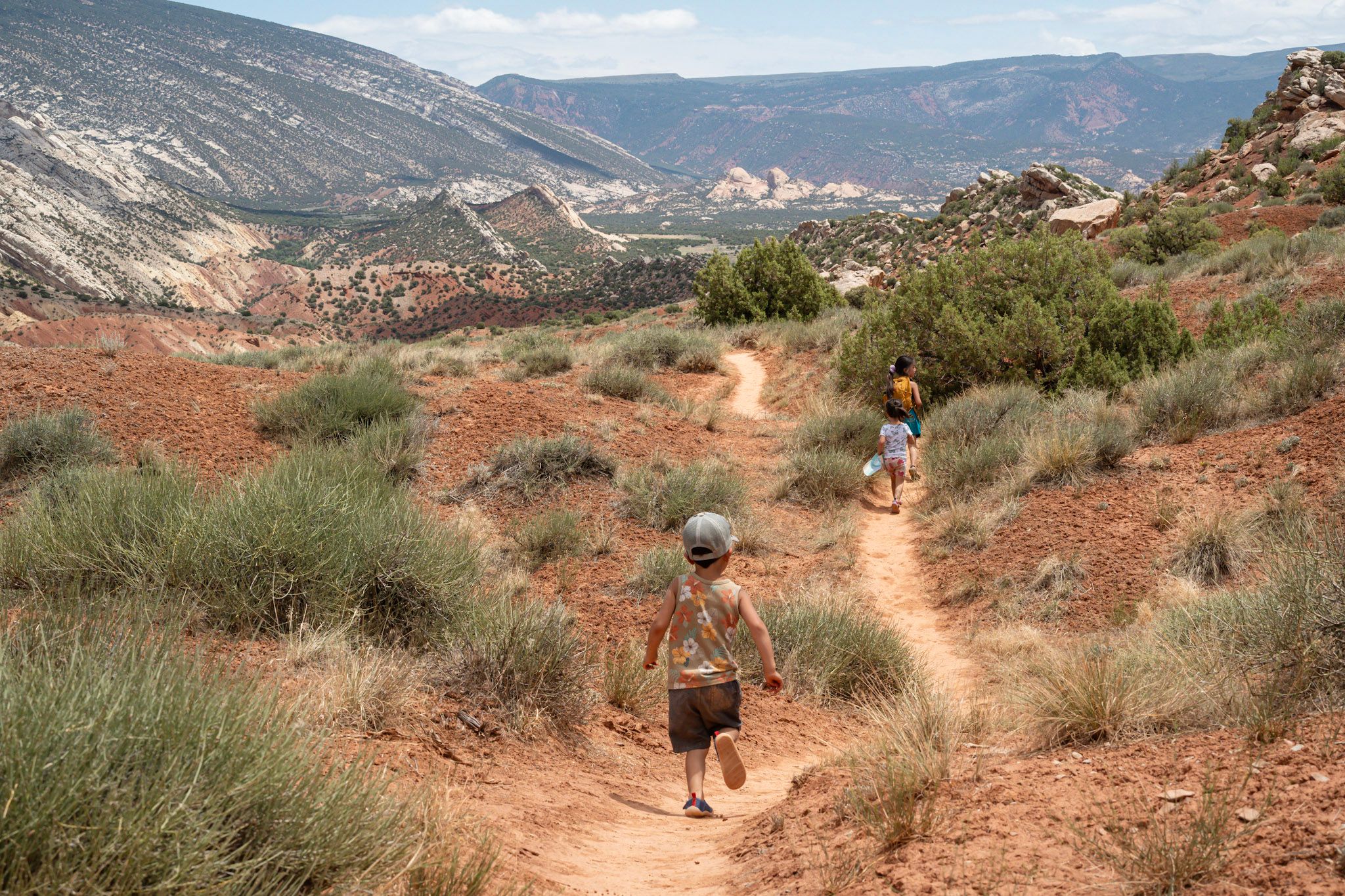 Two hikers walk along a red dirt trail through desert terrain with colorful cliffs in the distance. utah dinosaur national monument adventur