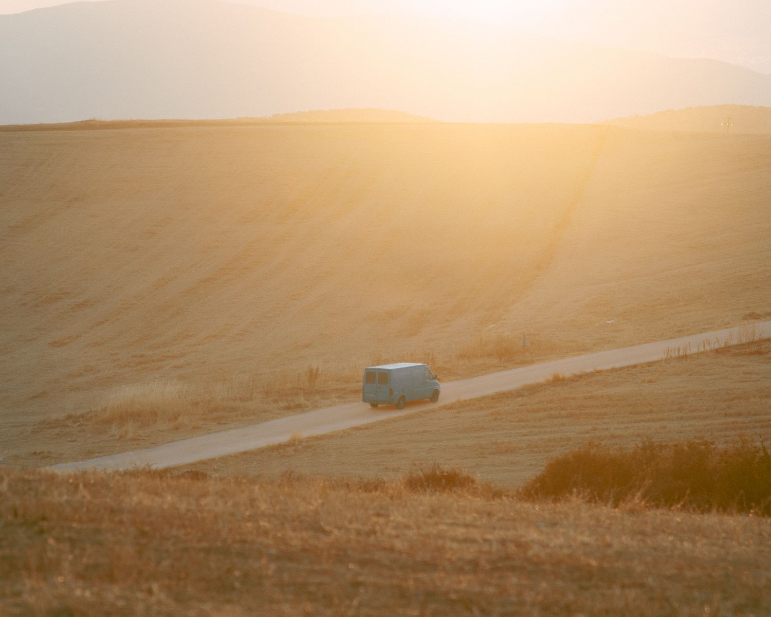 Car driving on a dirt road through open fields at sunset, viewed from a distance.