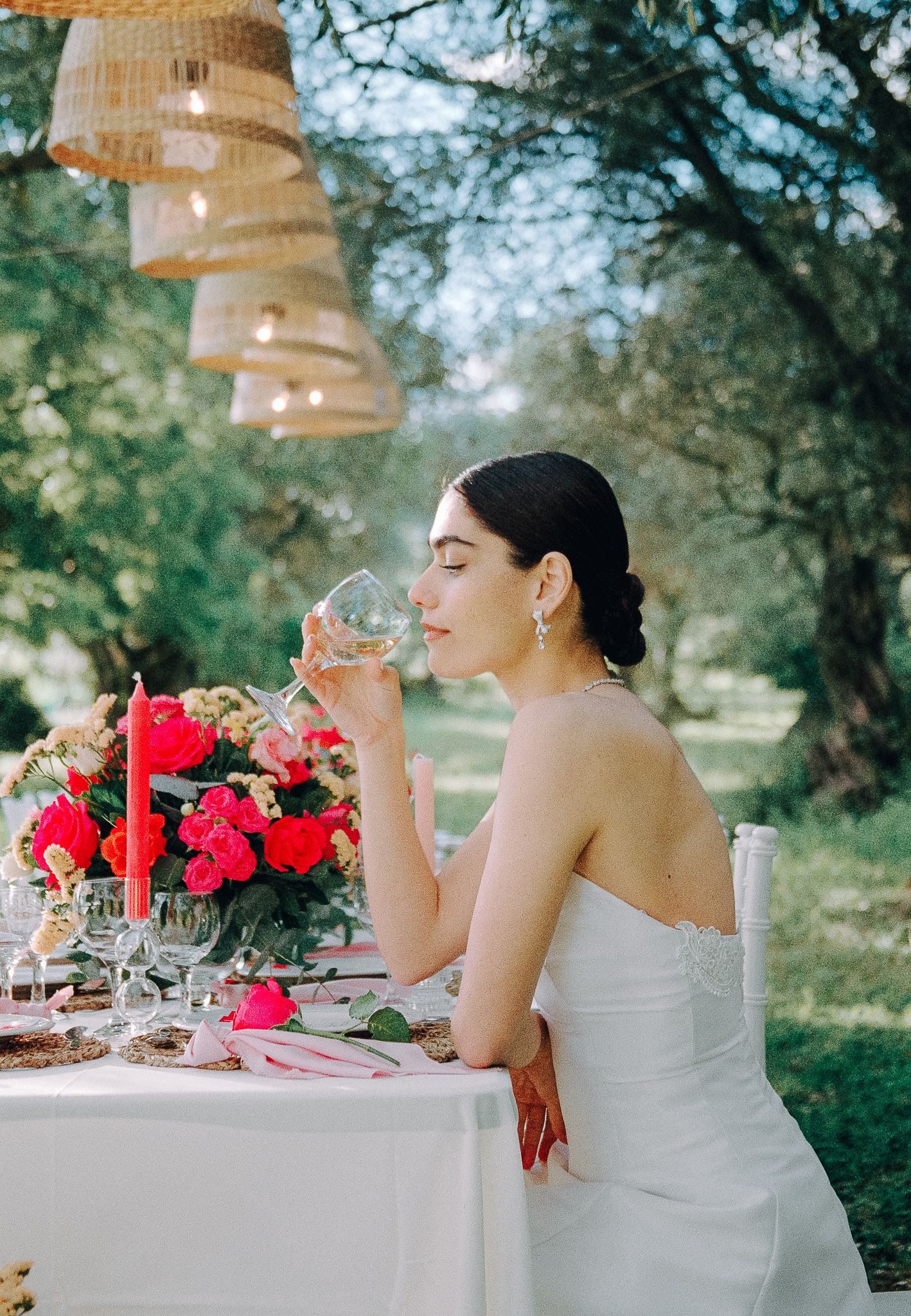 Bride in white dress sipping water at a styled outdoor table under olive trees.