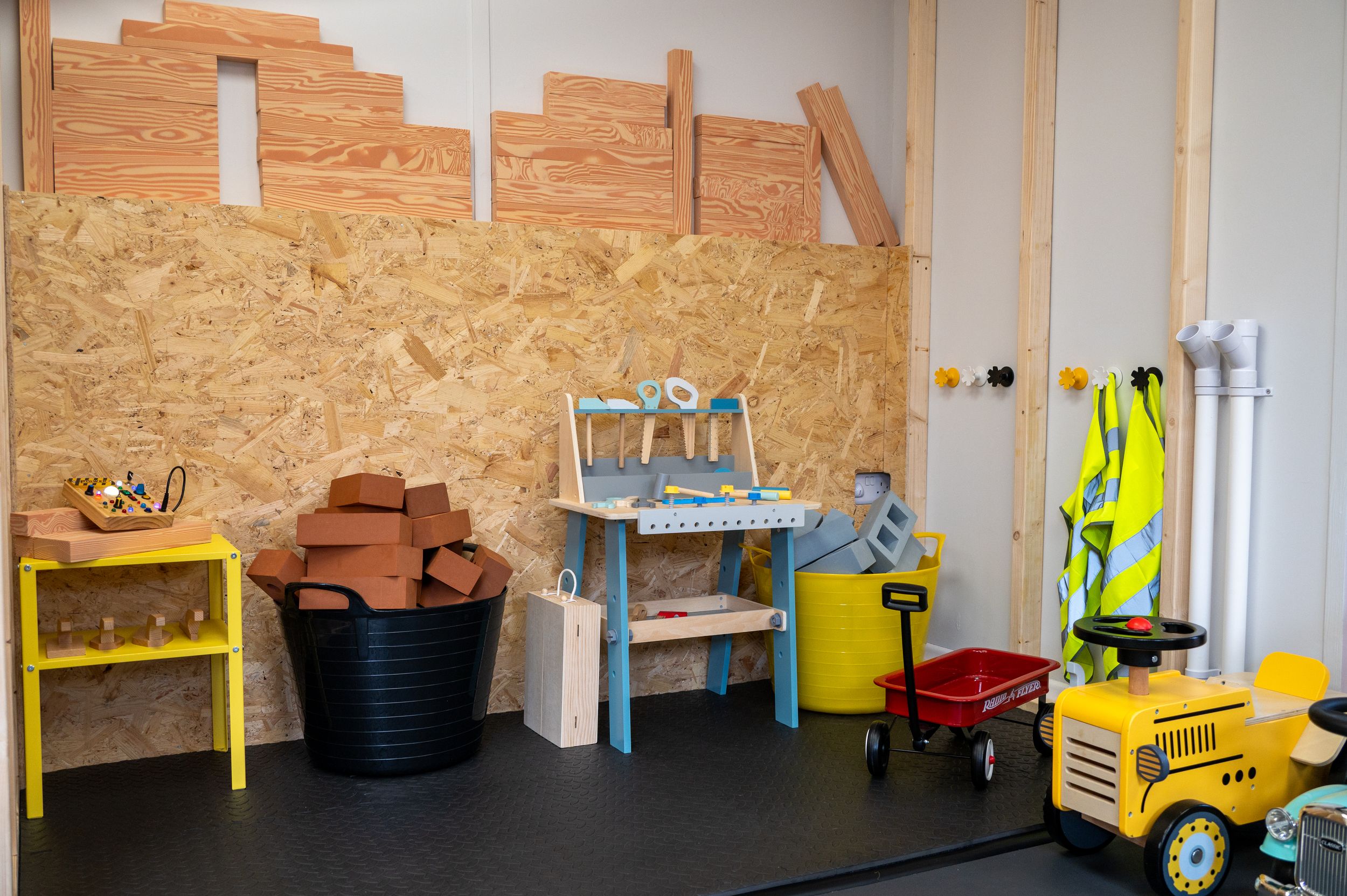 A children's play area with toy construction equipment, work bench, and high-visibility gear against a wooden wall.
