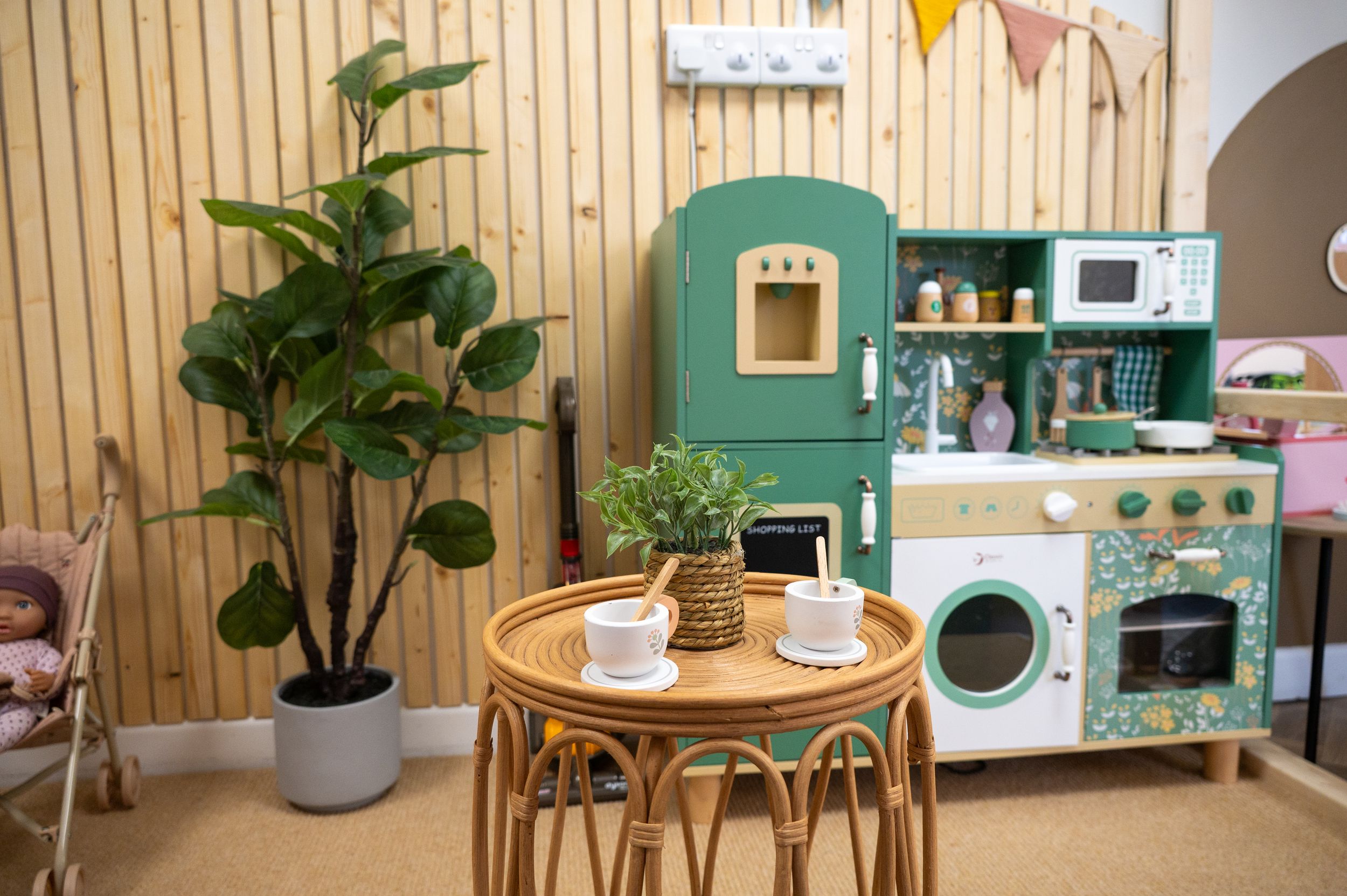 Cozy playroom corner with mint green toy kitchen, rattan table set, and large potted plants against wood paneled wall.
