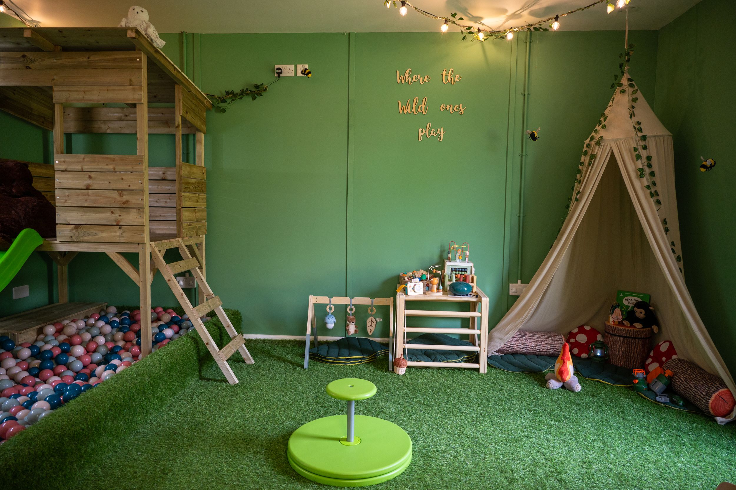 Fun playroom interior with green walls, artificial turf flooring, ball pit, climbing structure and teepee tent for children.