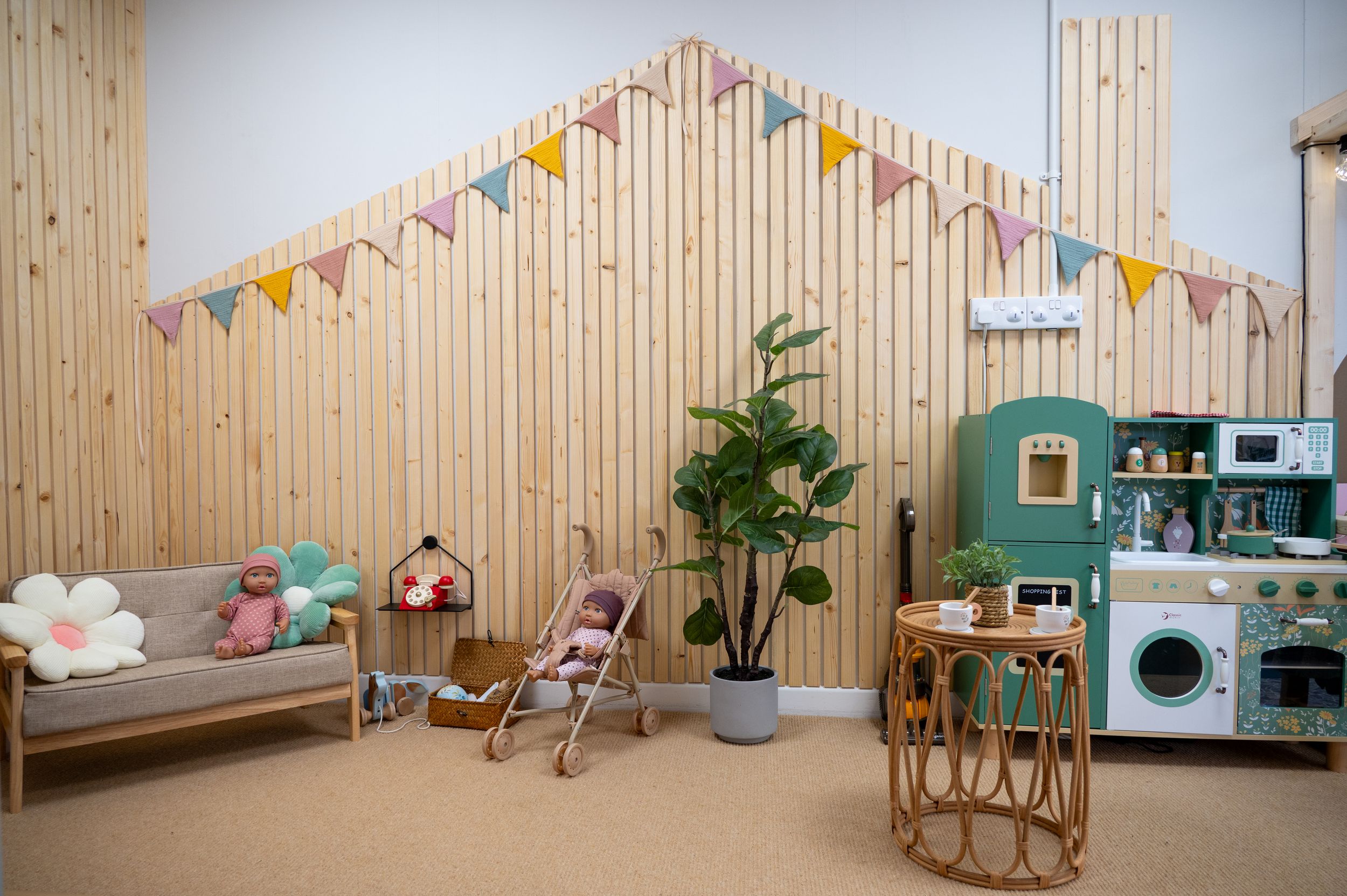 Modern playroom with wooden wall panels, colorful bunting flags, rattan furniture, and a toy kitchen set against teal accents.