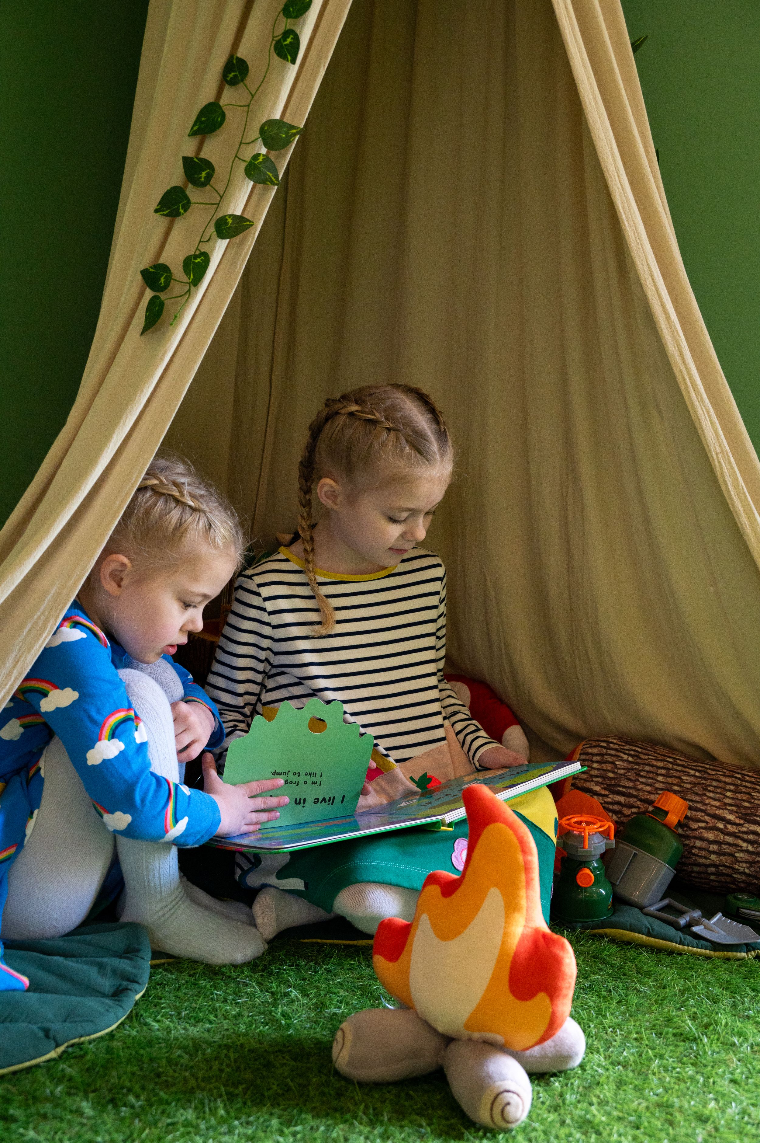 Two children reading books together in a cozy tent with green decorations and soft plush toys.