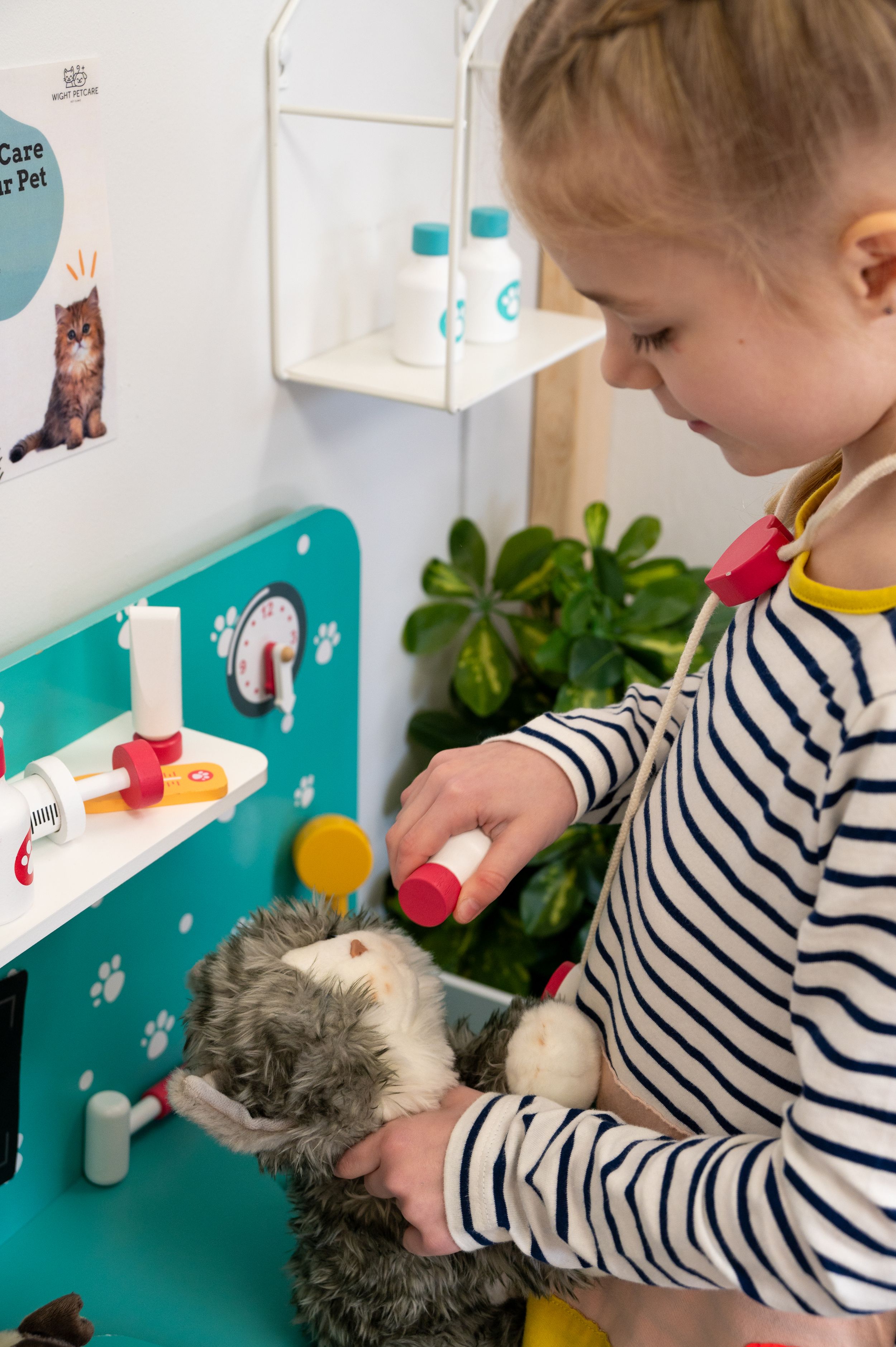A child in a striped shirt plays with a toy veterinary set while holding a stuffed gray cat toy at a play station.