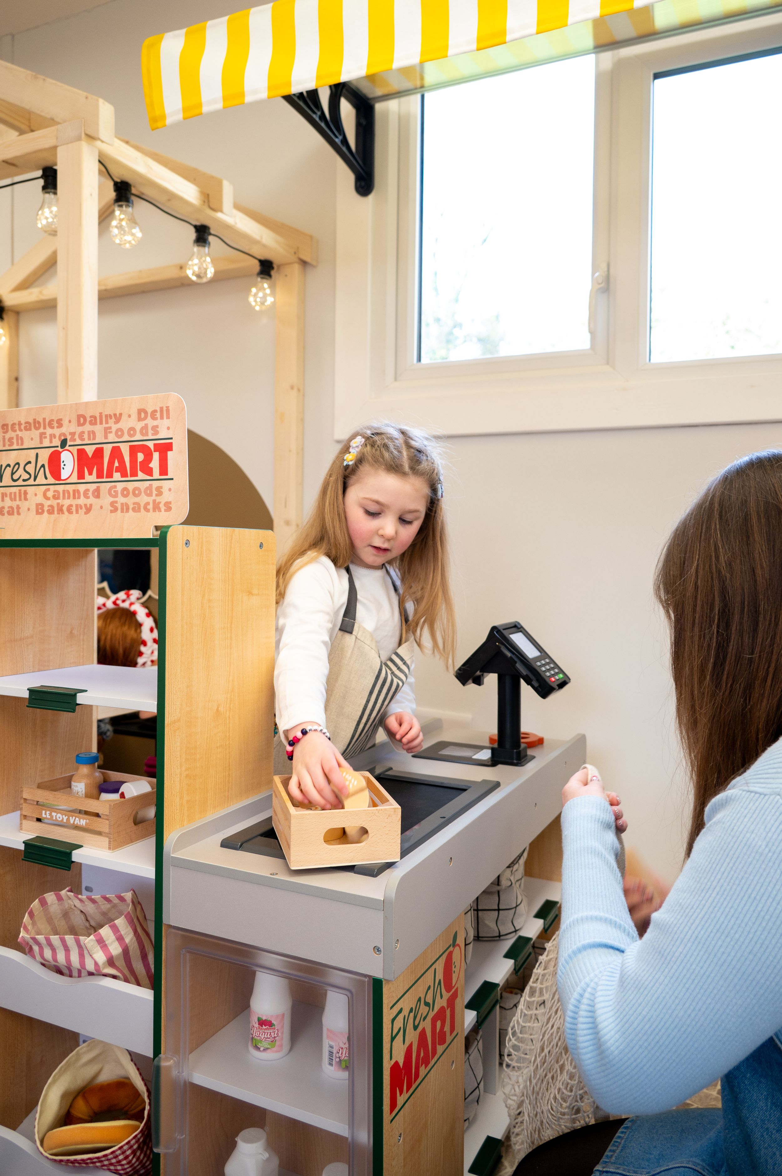 Children play at a pretend grocery store checkout with a wooden cash register and toy mart setup indoors.