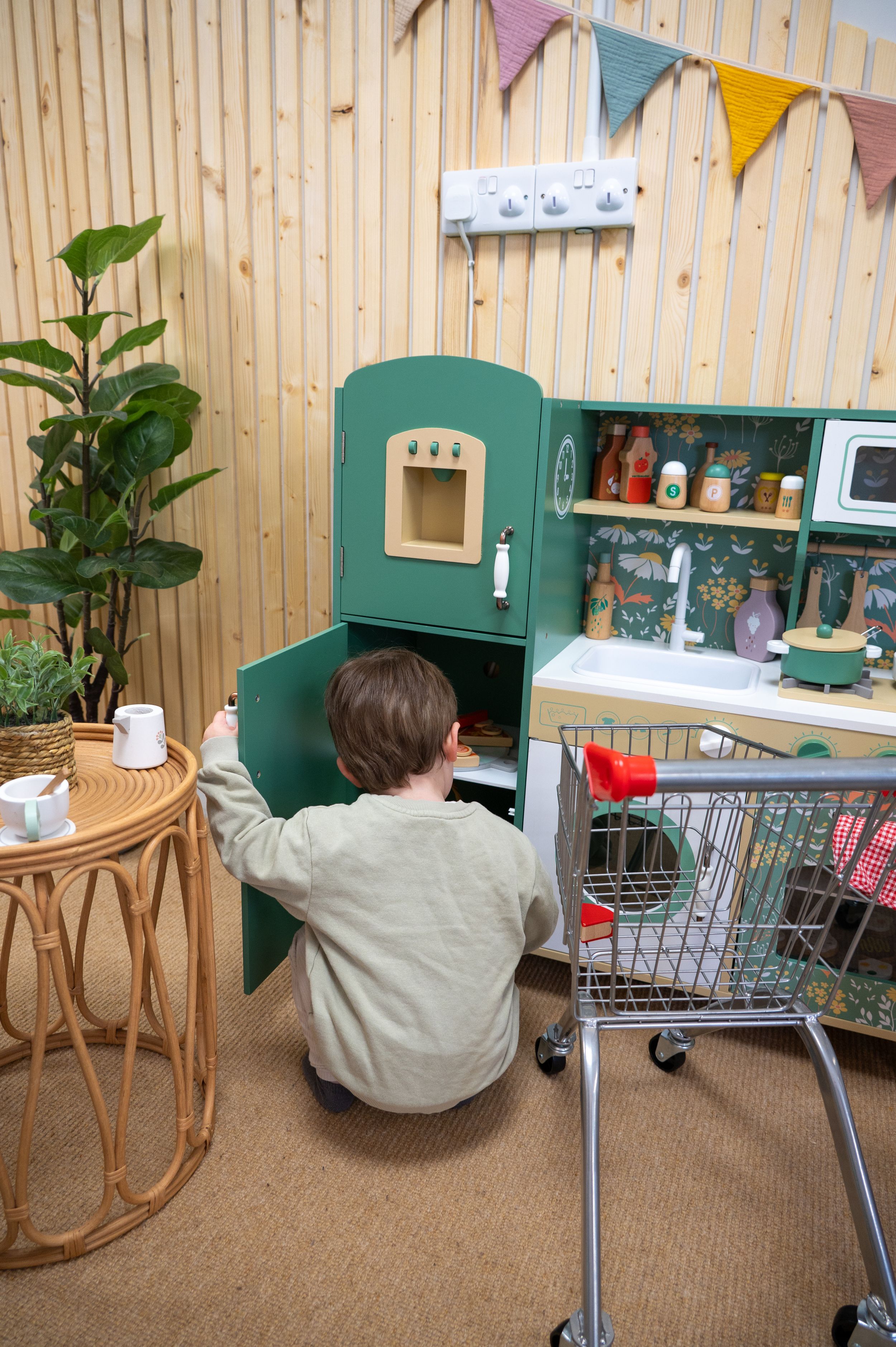 Child playing with toy kitchen setup featuring green play fridge, shopping cart and decorative bunting in playroom.