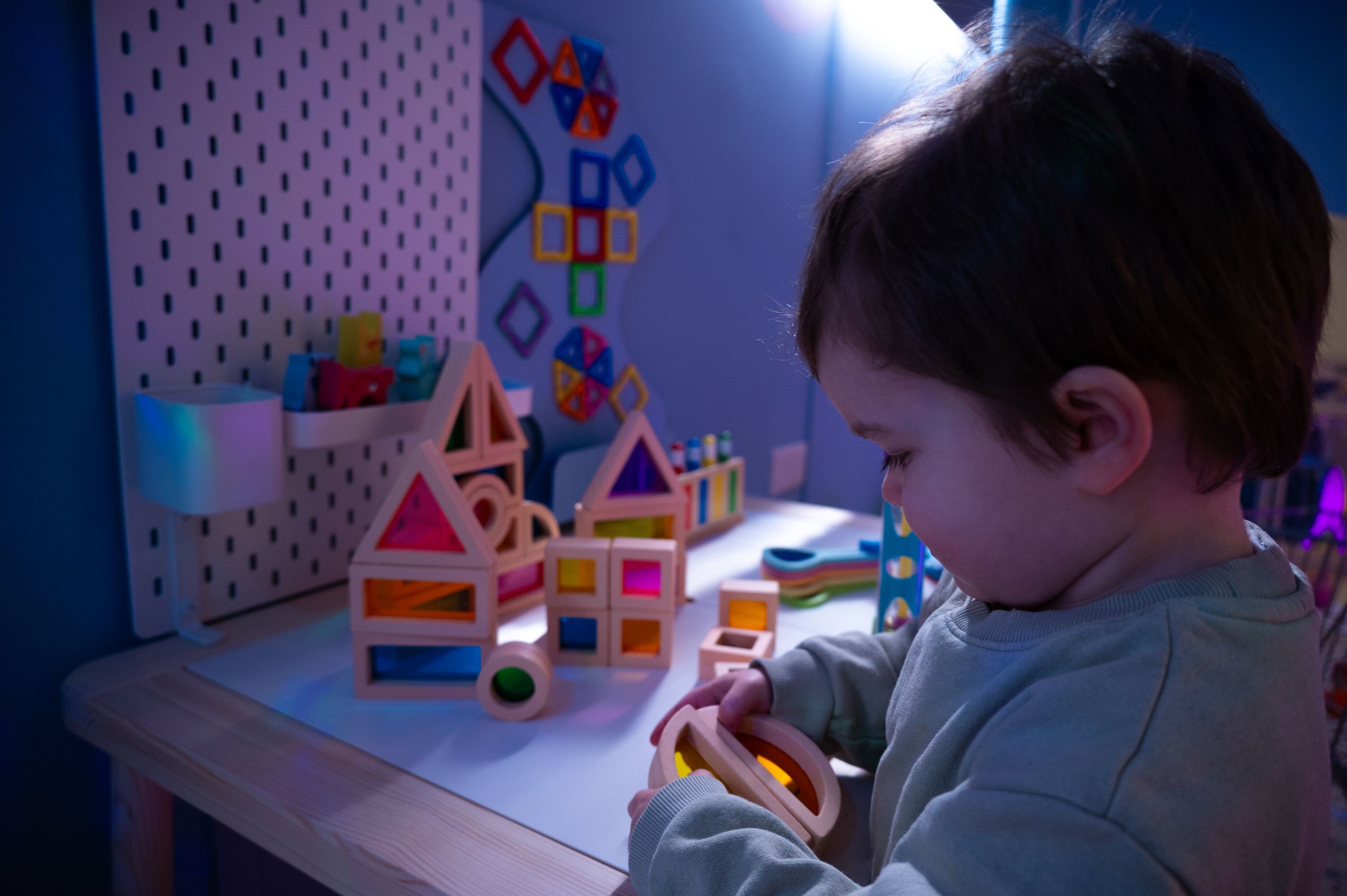 A young child plays with colorful geometric blocks and toys on a desk illuminated by blue and pink lighting.