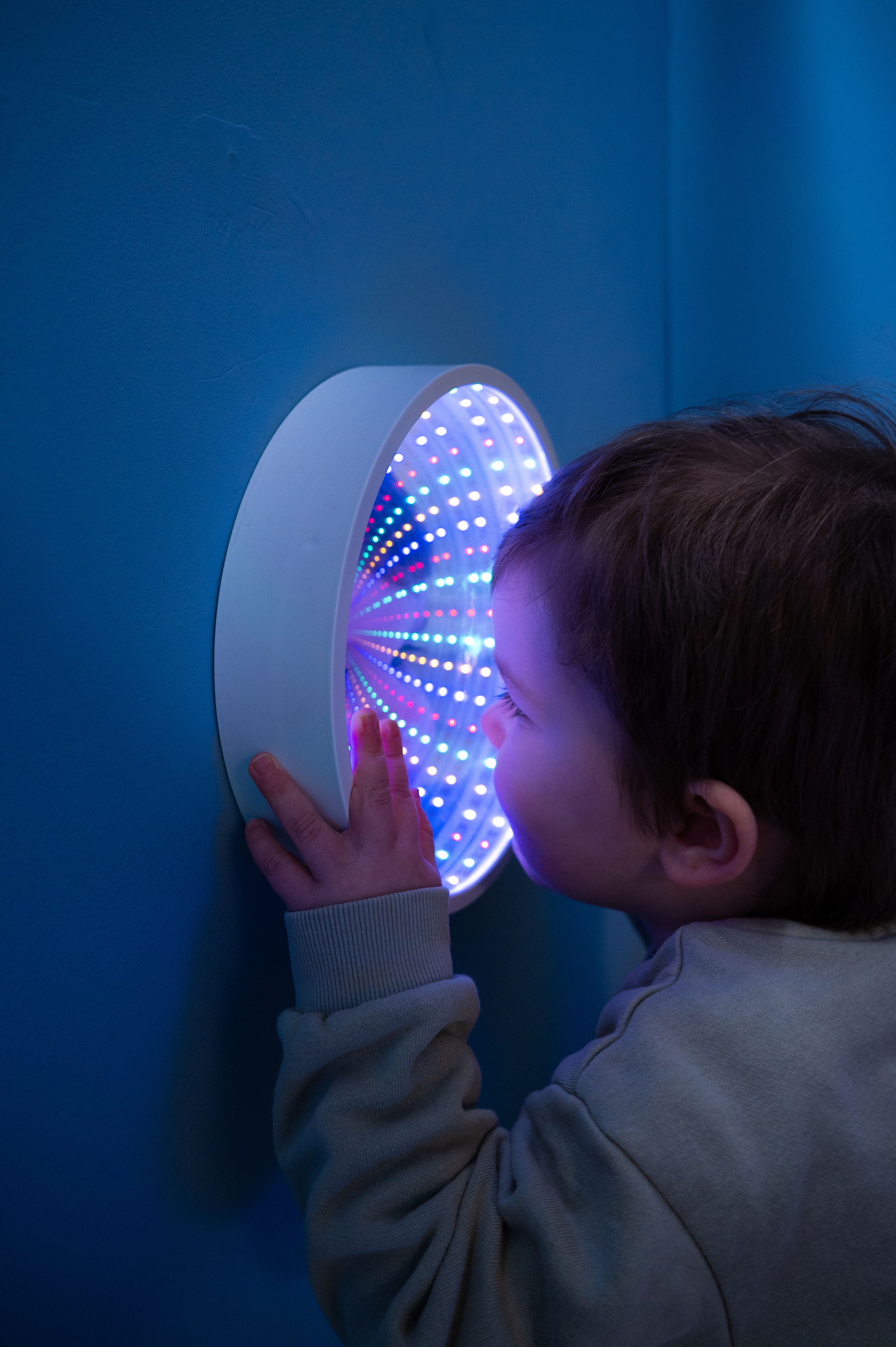 LED infinity mirror display emits colorful lights in dark room as small figure peers into glowing circular wall fixture.