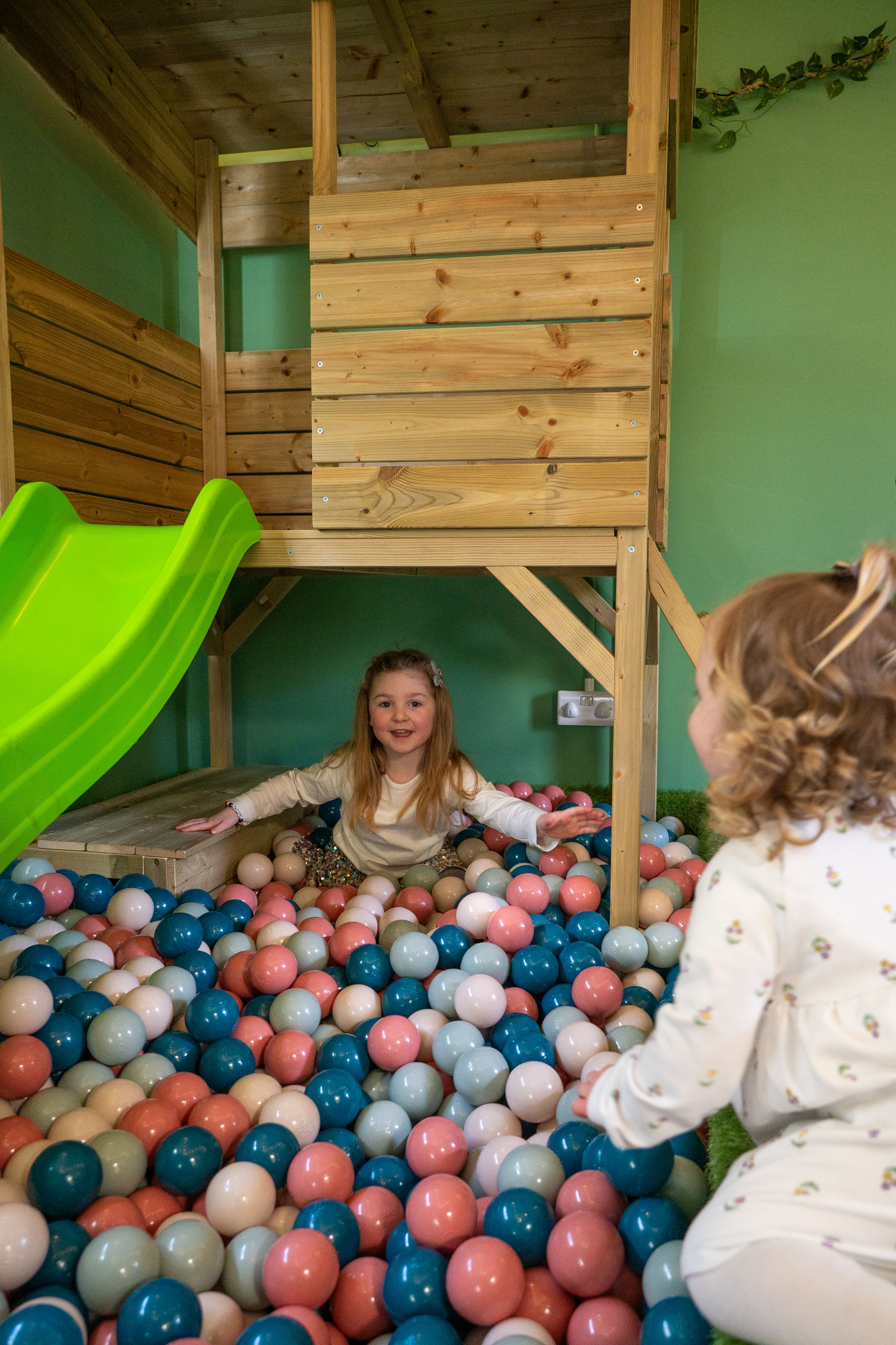 Children playing in a colorful ball pit beneath a wooden loft with green slide in playroom.