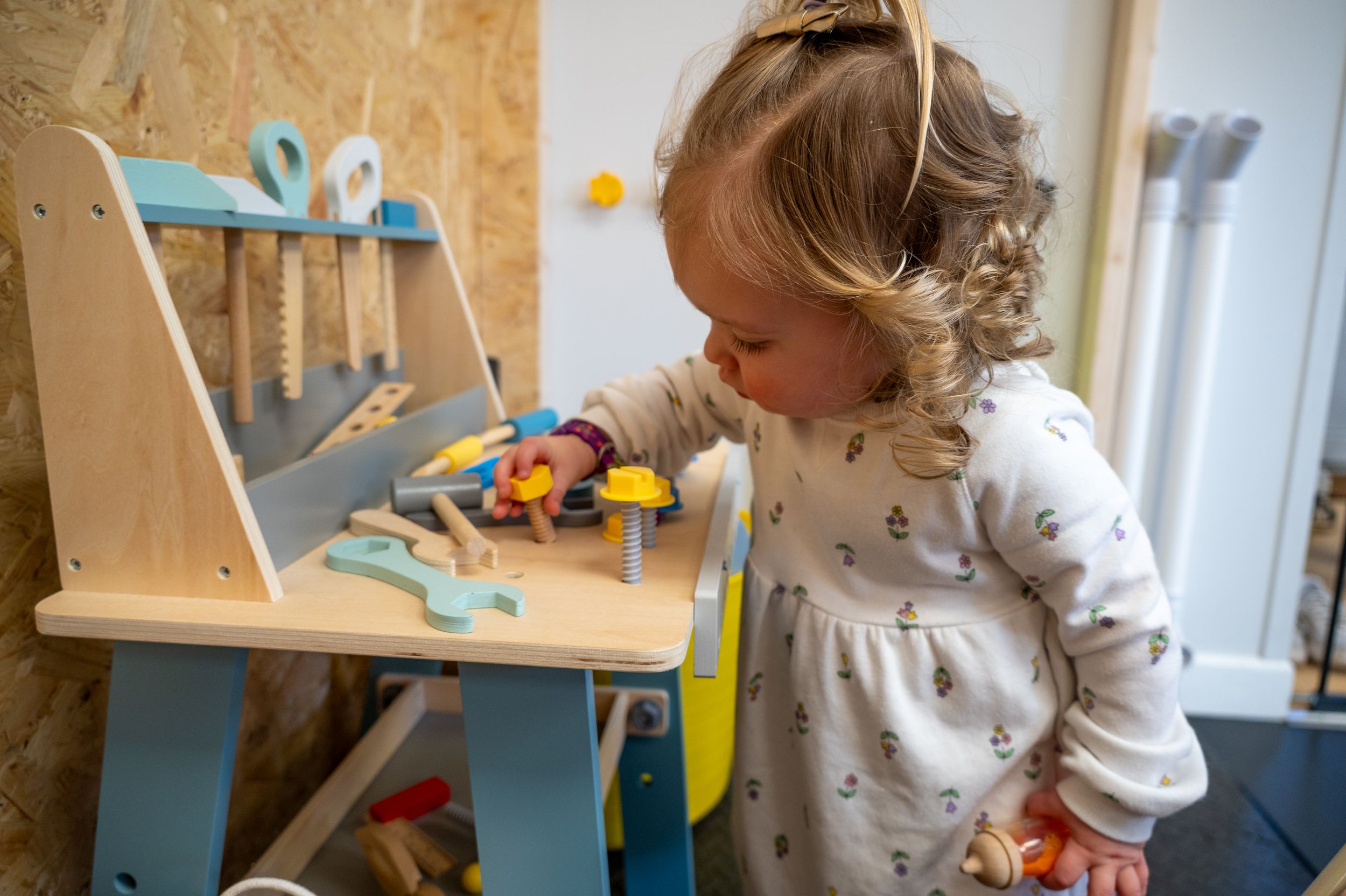 A small child wearing a white shirt plays at a wooden workbench with colorful toy tools and accessories.