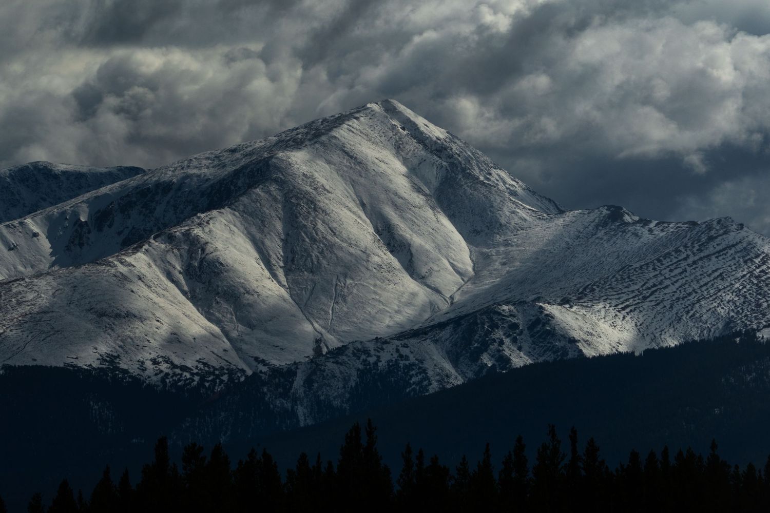 Snow-capped mountain peak under dramatic stormy clouds with dark forest silhouettes in the foreground.