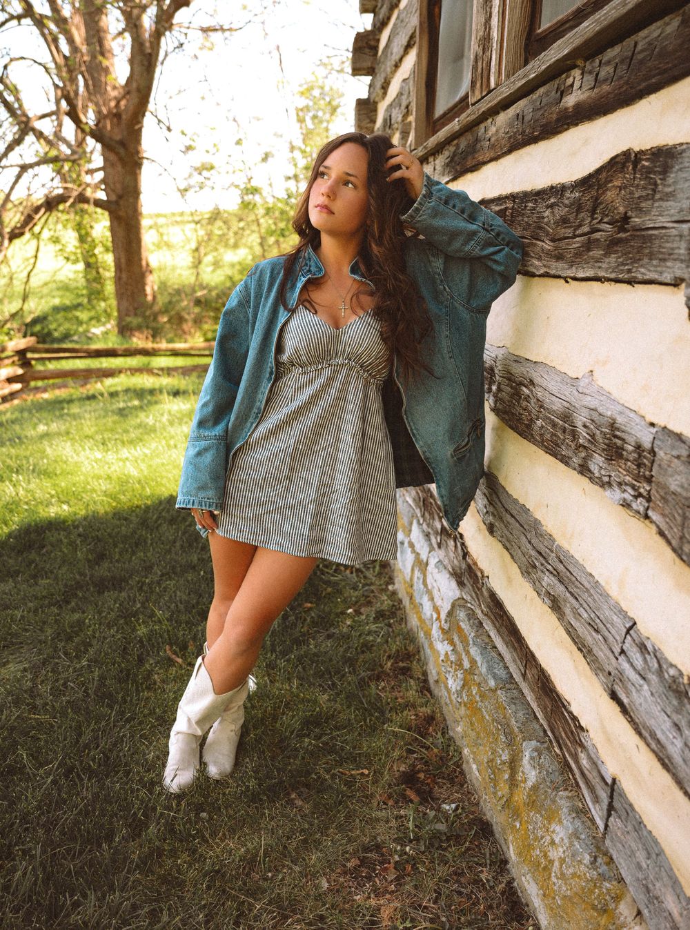 Young woman in denim jacket, gray mini dress, and white cowboy boots leaning against a rustic log cabin wall.