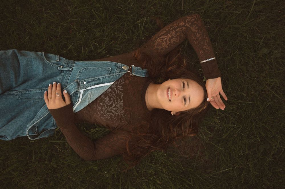 Young woman with long auburn hair lying on green grass, smiling, wearing denim overalls and a brown lace top.