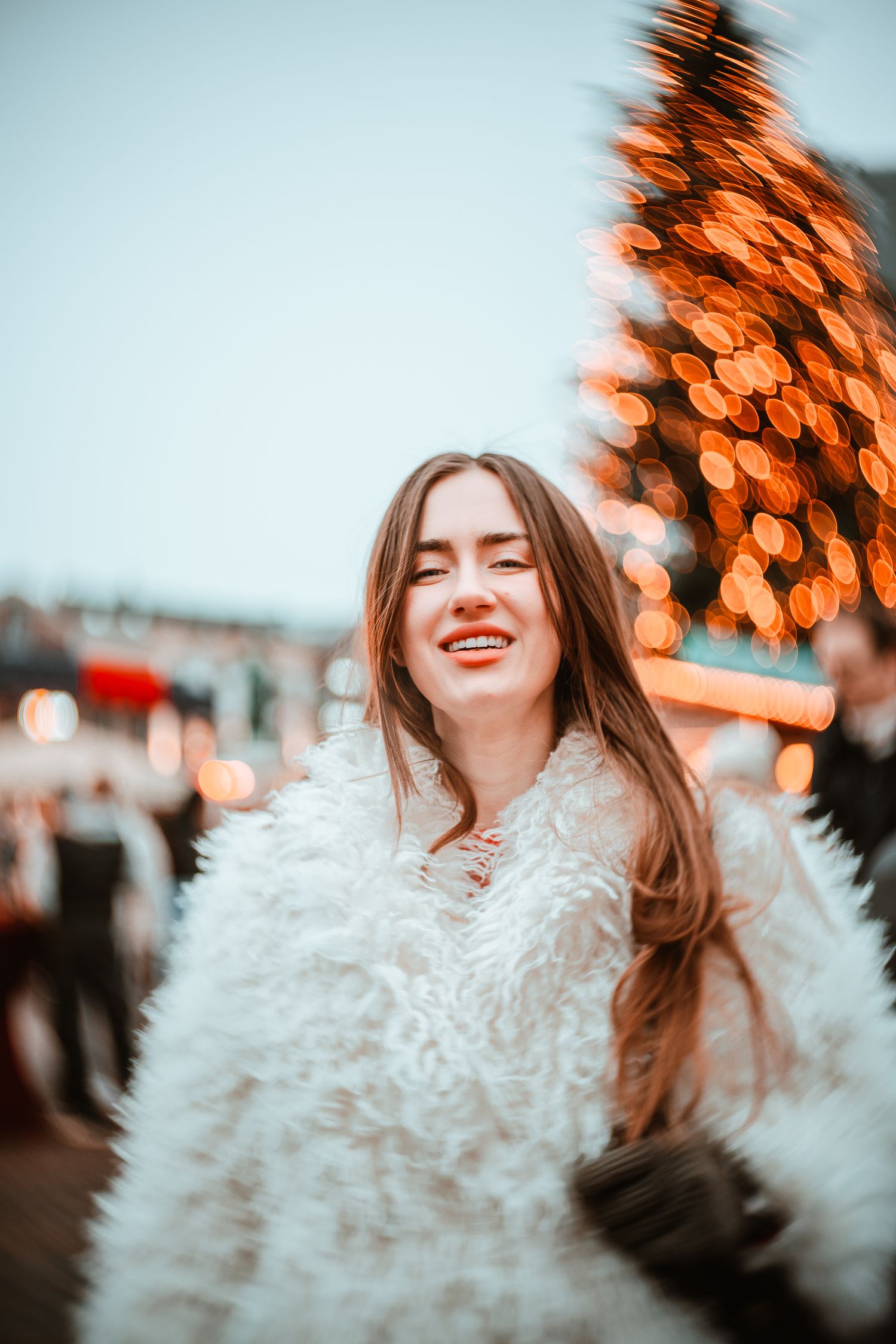 Woman withboyful expression and white fuzzy coat captured against warm orange bokeh lights in winter setting. Shot with New Petzval lens.