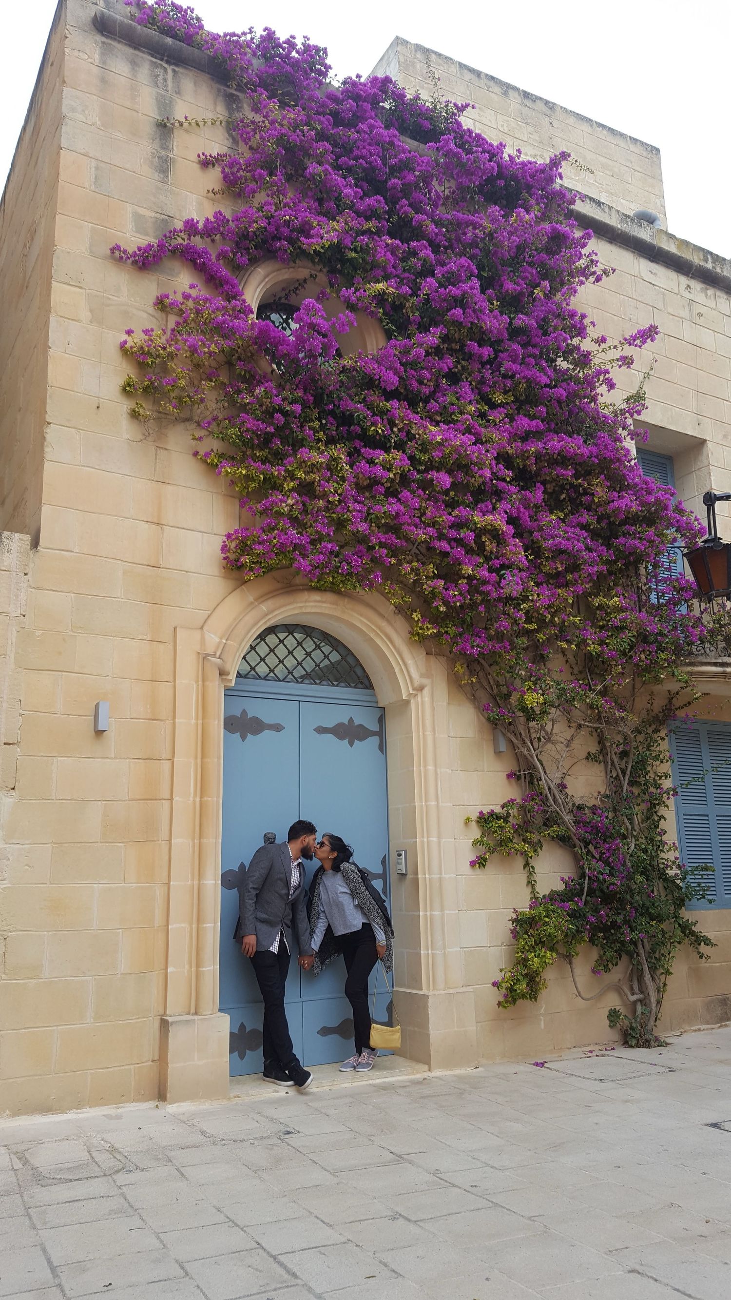 Beautiful purple bougainvillea flowers draping over a beige stone building with a blue door.
