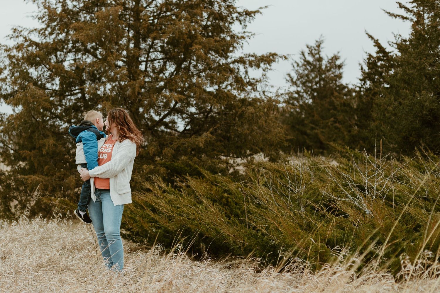 A parent holding a child stands in a field of tall grass with evergreen trees in the background.