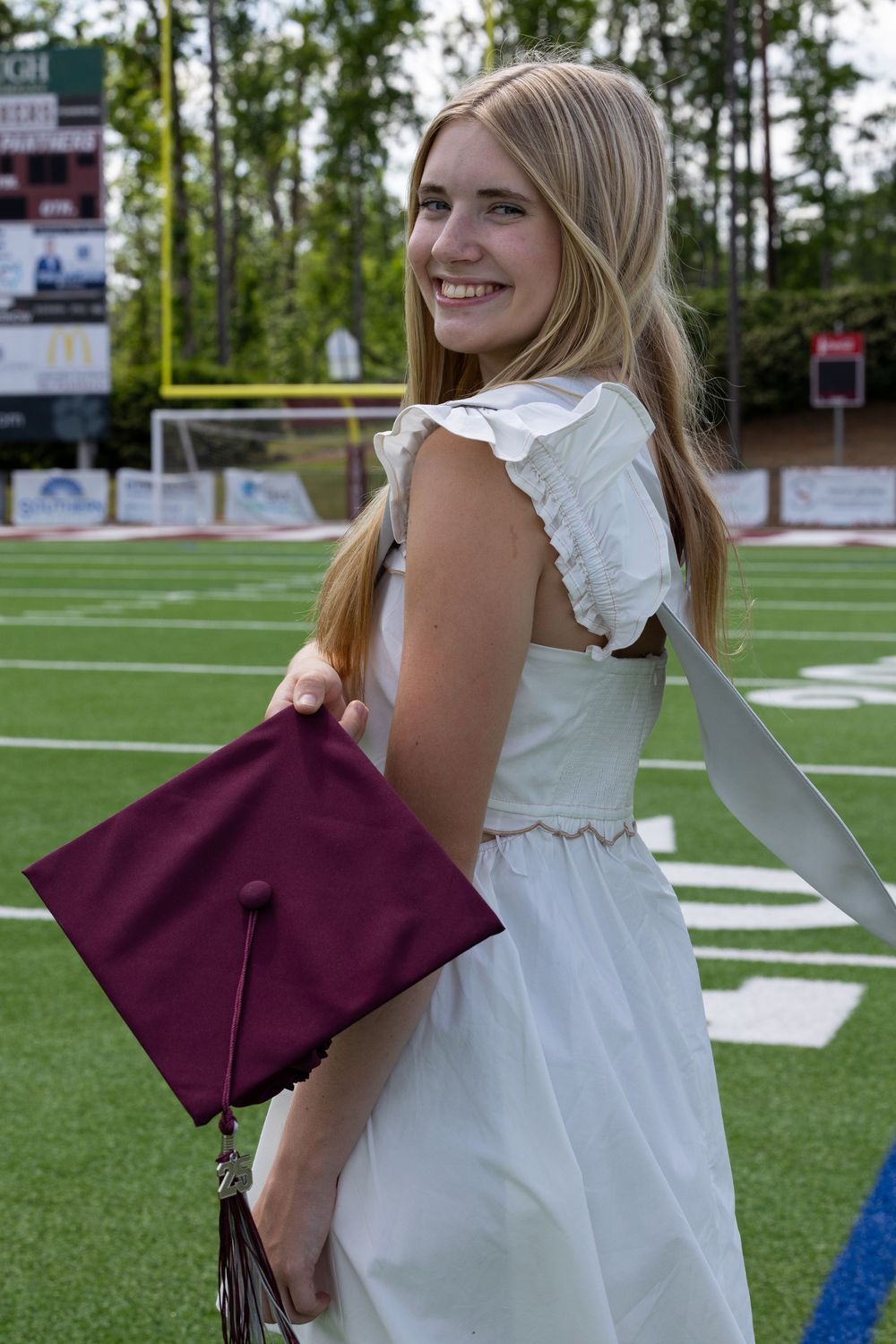 Graduate in white dress holds burgundy graduation cap while posing on football field with trees in background.