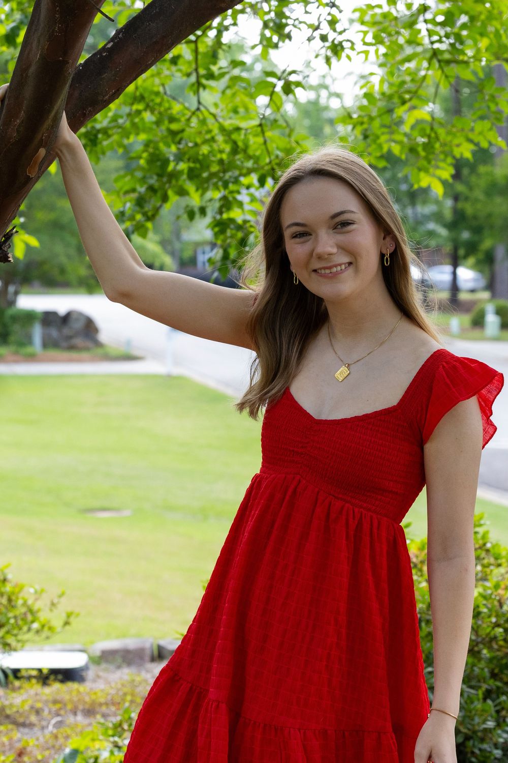 A person in a bright red summer dress poses outdoors near a tree with green foliage and a garden in the background.