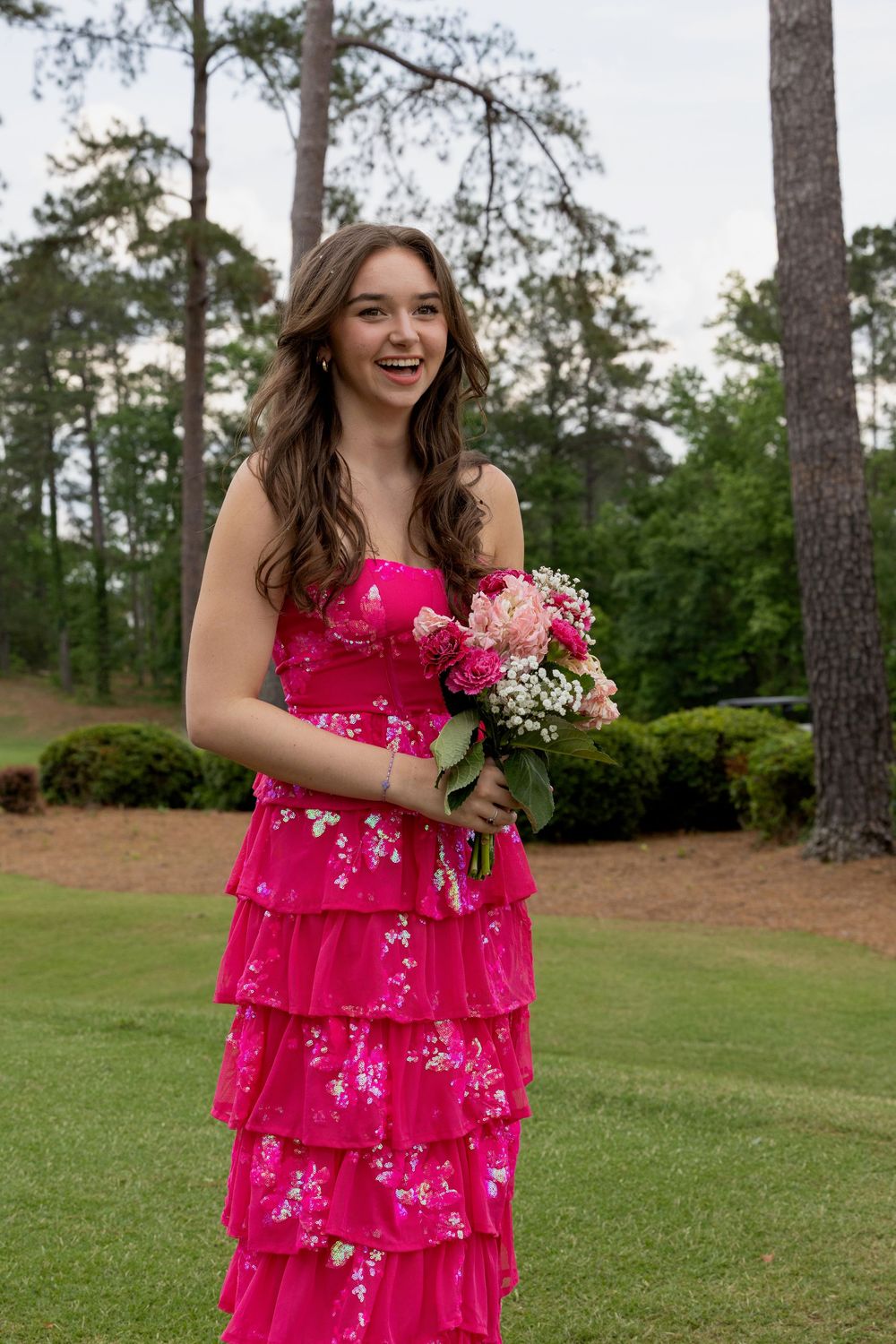 Person in bright pink tiered floral dress holds pink and white bouquet while standing on manicured lawn near pine trees.