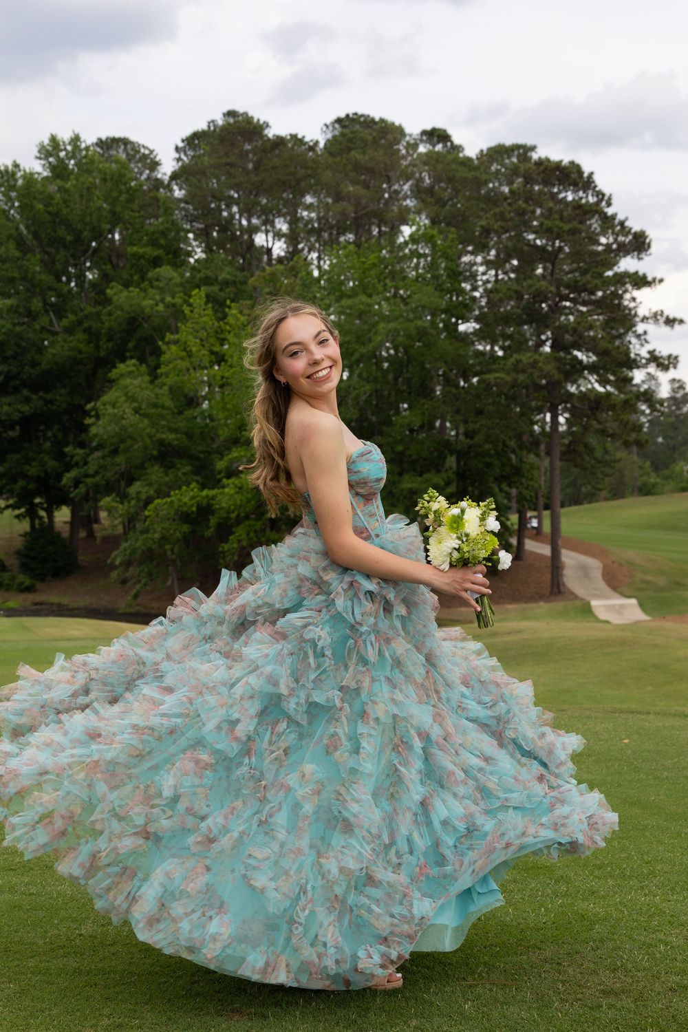Someone twirls in an elegant turquoise and white ruffled ballgown while holding a bouquet on a green golf course lawn.
