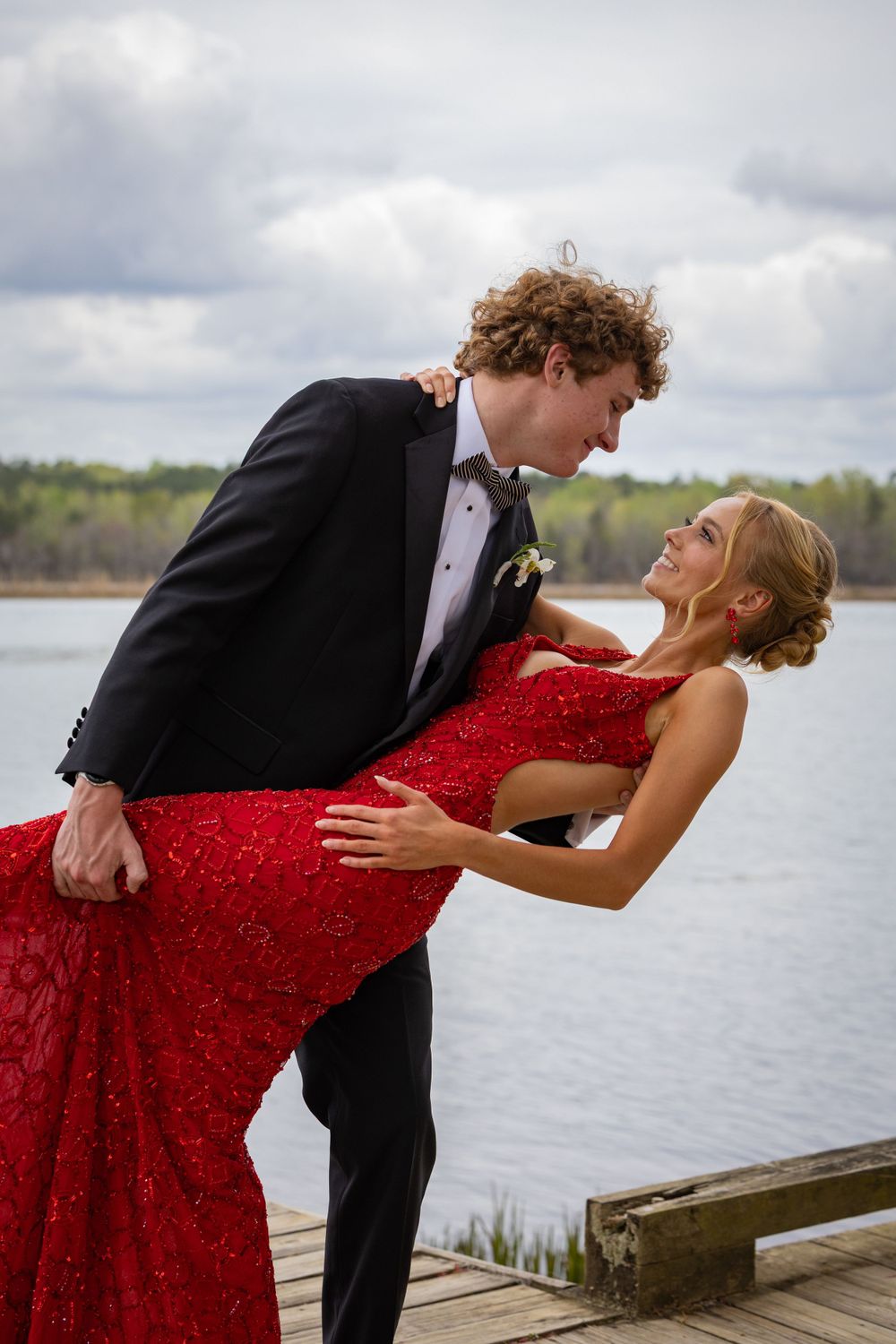 Couple in formal attire shares romantic dip pose on dock by lake.