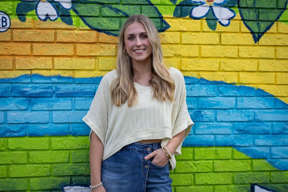 A person in casual wear poses against a vibrant yellow and blue painted brick wall.